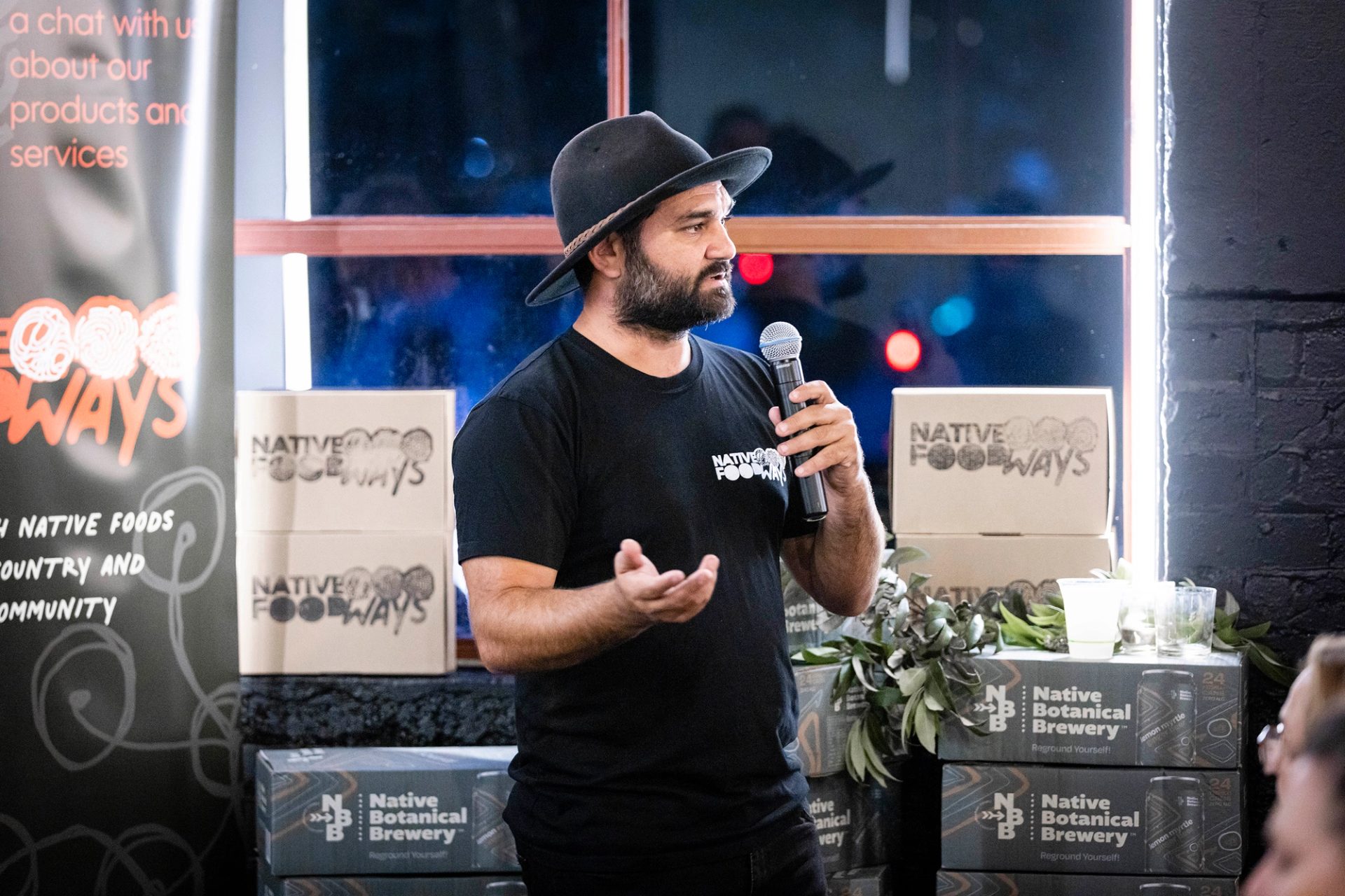 Man speaks on microphone to crowd, with boxes of Native Foodways and Native Botanical Brewery around them