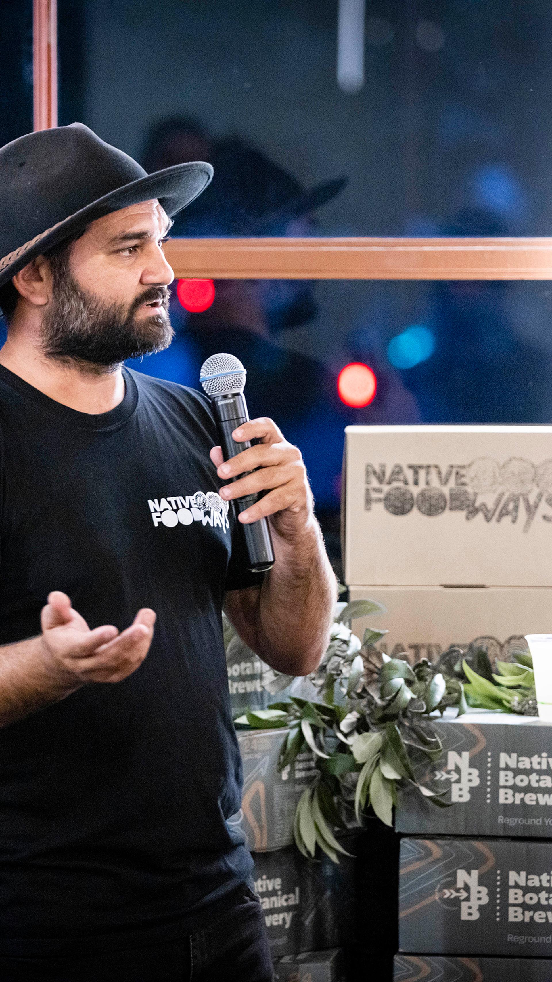 Man speaks on microphone to crowd, with boxes of Native Foodways and Native Botanical Brewery around them