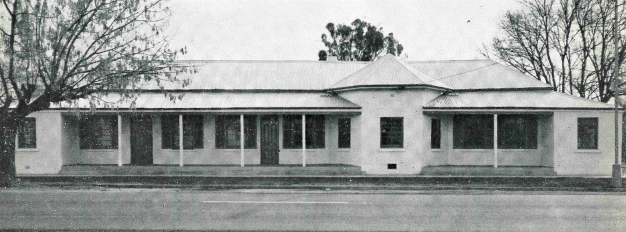 Black and white photograph of a one-storey museum exterior with white walls and a corrugated iron roof