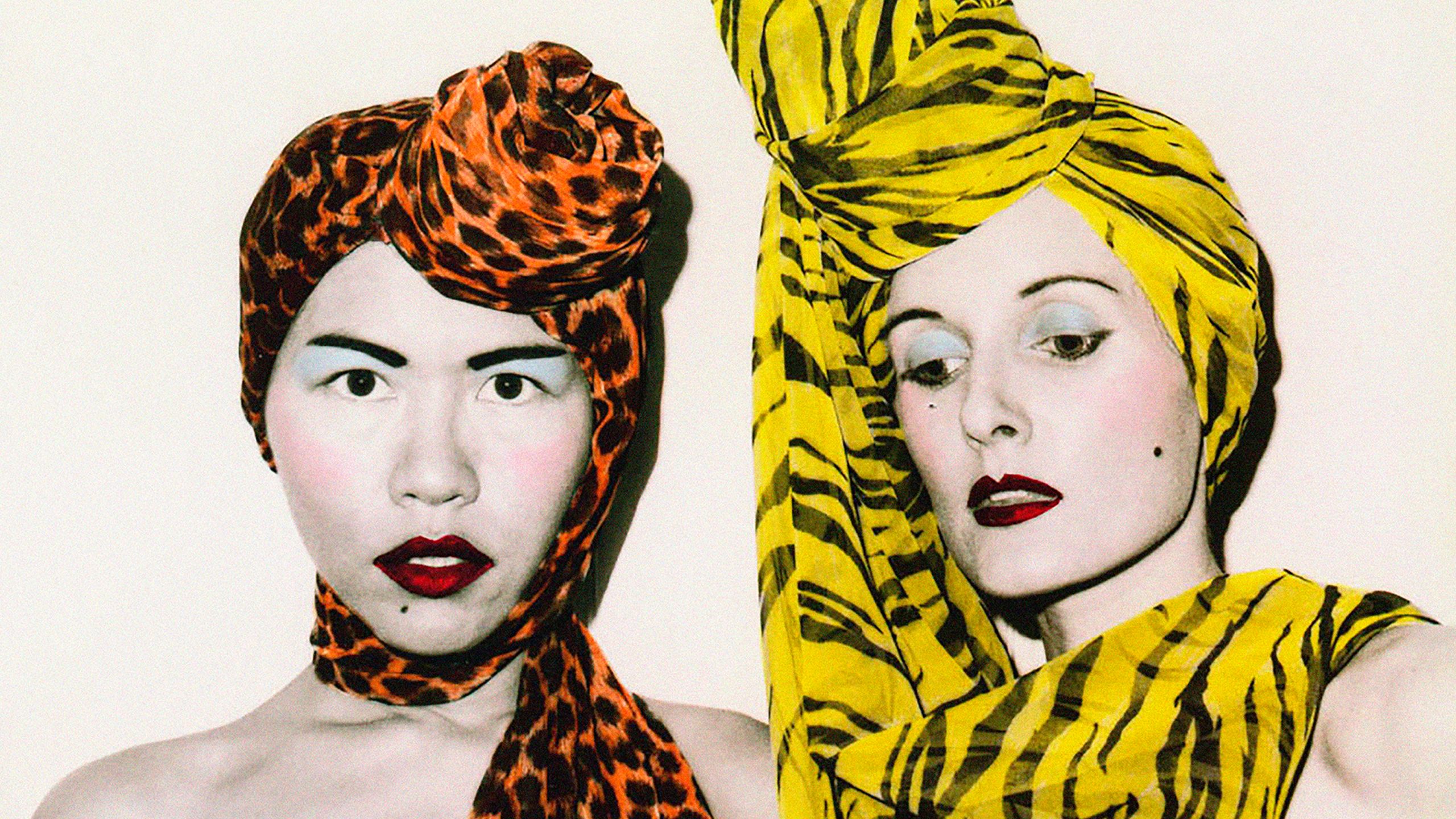 A photograph of fashion designers Jenny Lee and Linda Jackson. Both wear elaborate and highly colored headscarfs, along with a full face of carefully applied make-up.