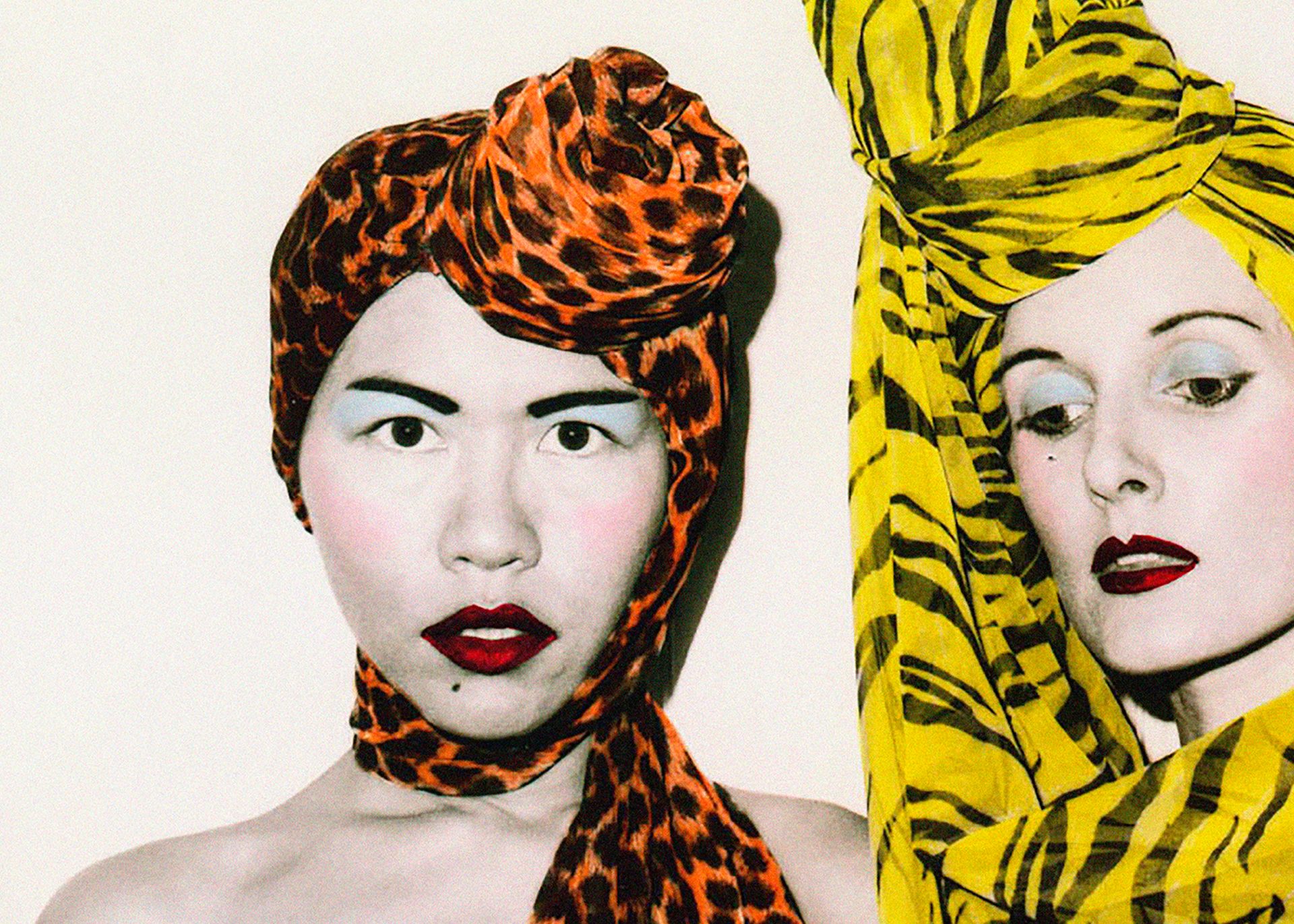 A photograph of fashion designers Jenny Lee and Linda Jackson. Both wear elaborate and highly colored headscarfs, along with a full face of carefully applied make-up.