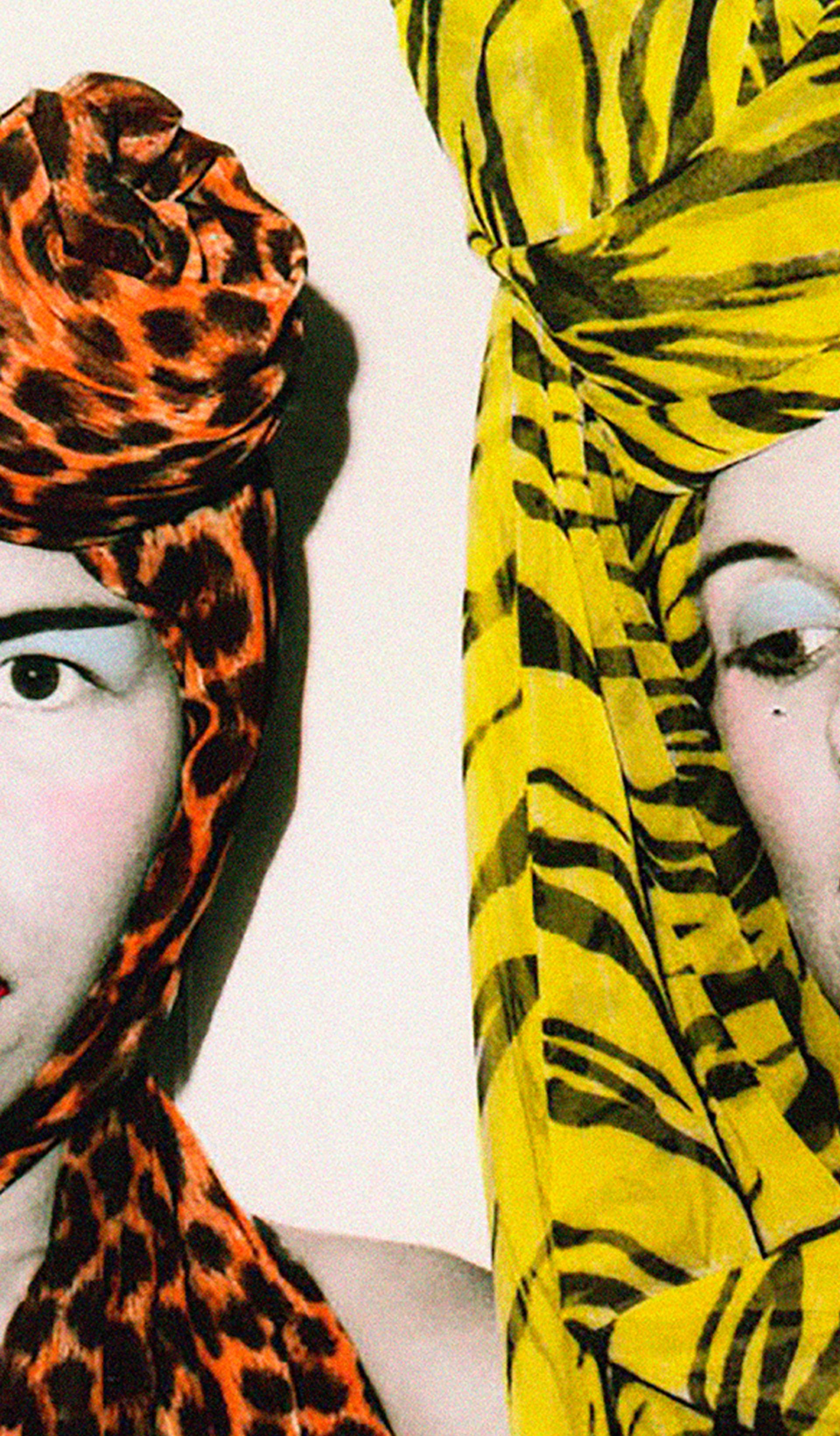 A photograph of fashion designers Jenny Lee and Linda Jackson. Both wear elaborate and highly colored headscarfs, along with a full face of carefully applied make-up.