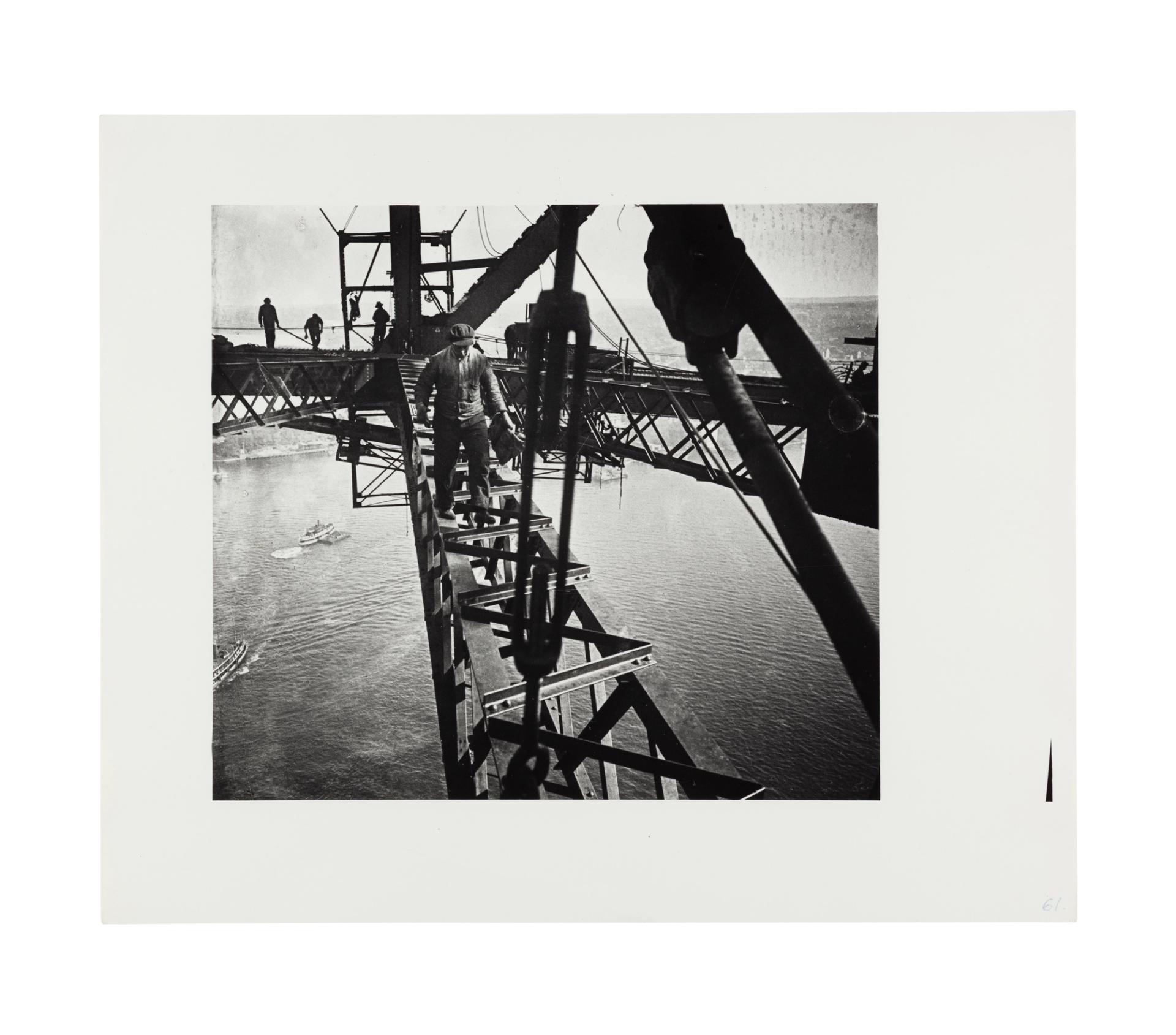 Image of a man walking in the direction of the camera across a beam of the harbour bridge. Three other figures are visible in the distance on an adjacent beam working. In the background the harbour and some ferries are visible through the gaps between the bridge beams.