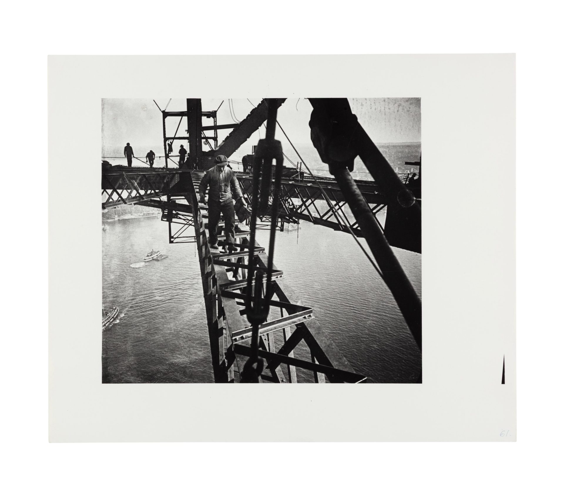 Image of a man walking in the direction of the camera across a beam of the harbour bridge. Three other figures are visible in the distance on an adjacent beam working. In the background the harbour and some ferries are visible through the gaps between the bridge beams.