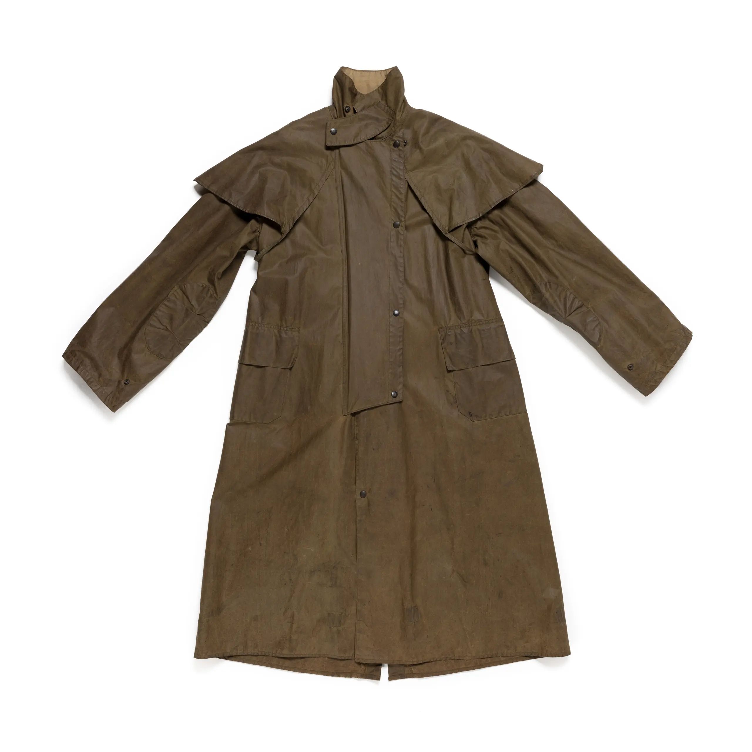 Brown Driza-Bone waterproof riding coat. Loose fitting coat to calf length with a small fold down collar which can be pulled up around neck and fastened with strap and snap closure. Short self fabric cape over shoulders, centre front opening fastening with snap closures and concealed beneath storm flap. Long sleeves with adjustable wrist snaps and elbow patches of same fabric. Two large patch pockets at front hip.