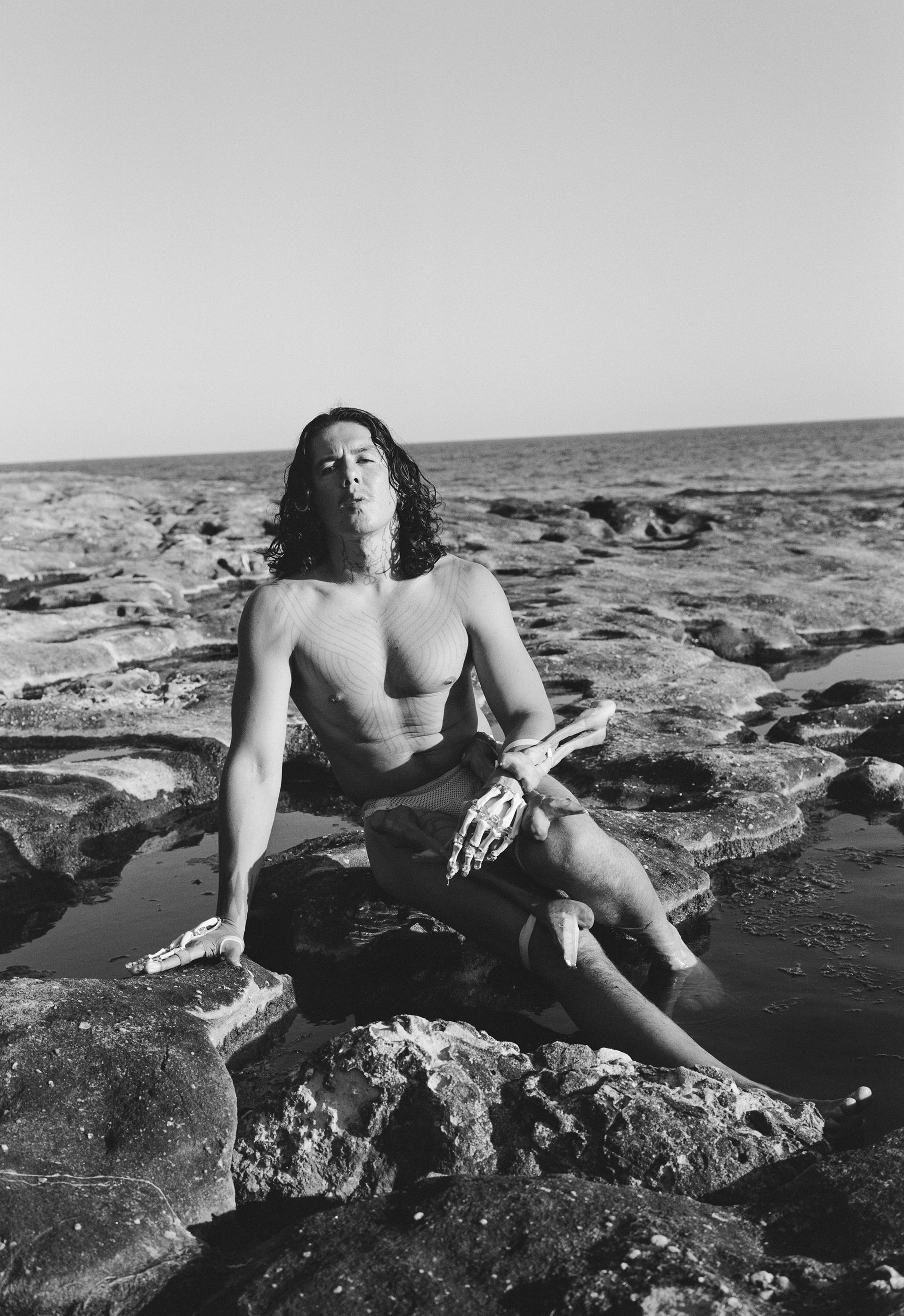 A black and white photograph of a man sitting on rocks at the beach. He is shirtless and wears a skeleton’s arm over his left arm.