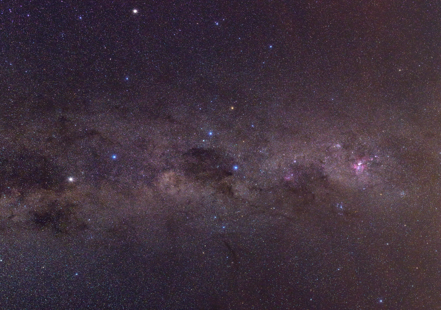 Colour photograph of the constellation of the Southern Cross and the Pointers, amongst a dense field of background stars that are blue, white and red. There are pink-ish regions (nebulae) and dark patches (interstellar dust clouds) visible too.