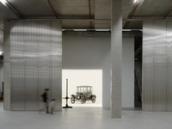 A tall and shorter human figures walk through the gigantic storage house, to find a car behind the wall in a white room.