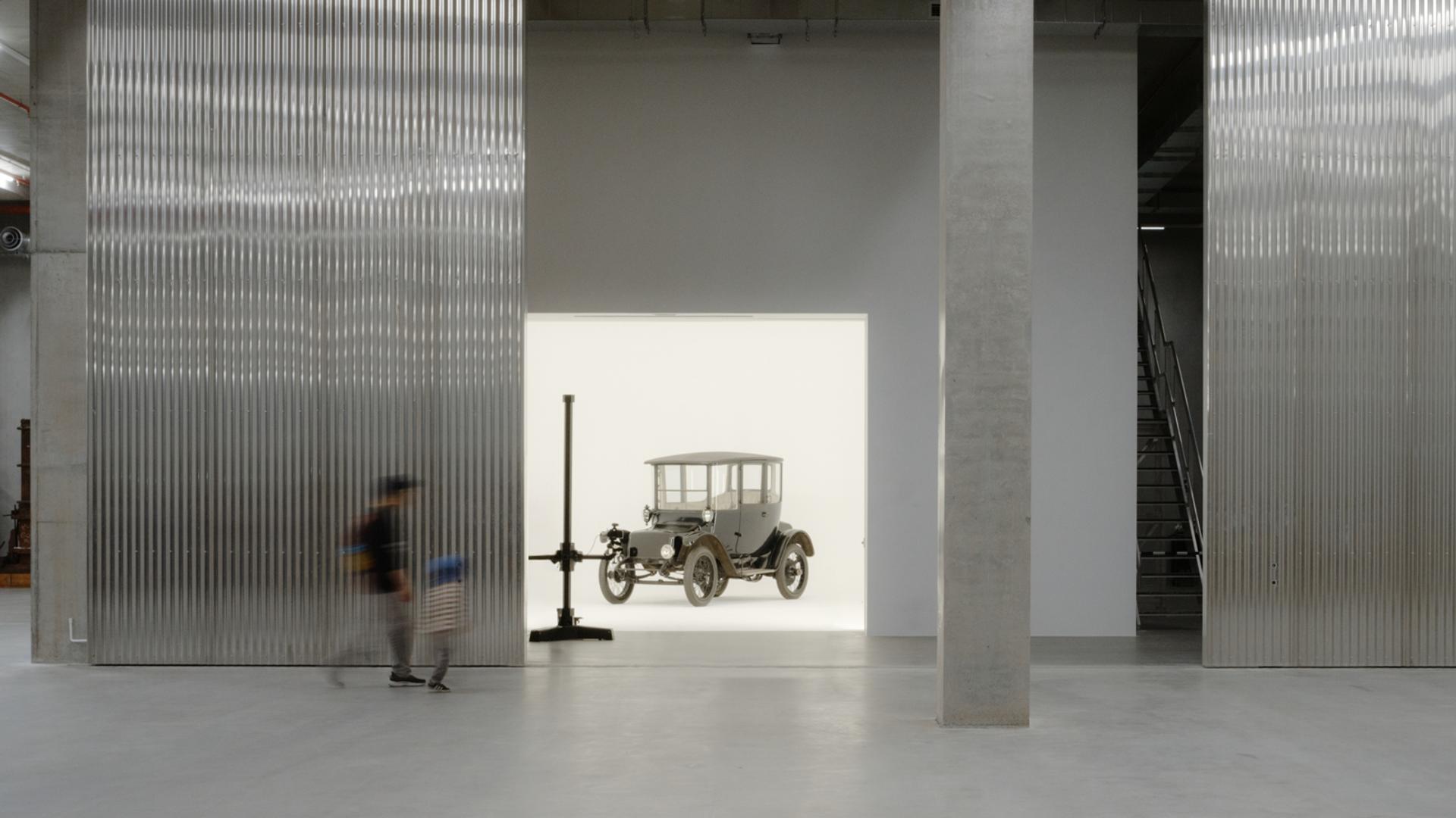A tall and shorter human figures walk through the gigantic storage house, to find a car behind the wall in a white room.