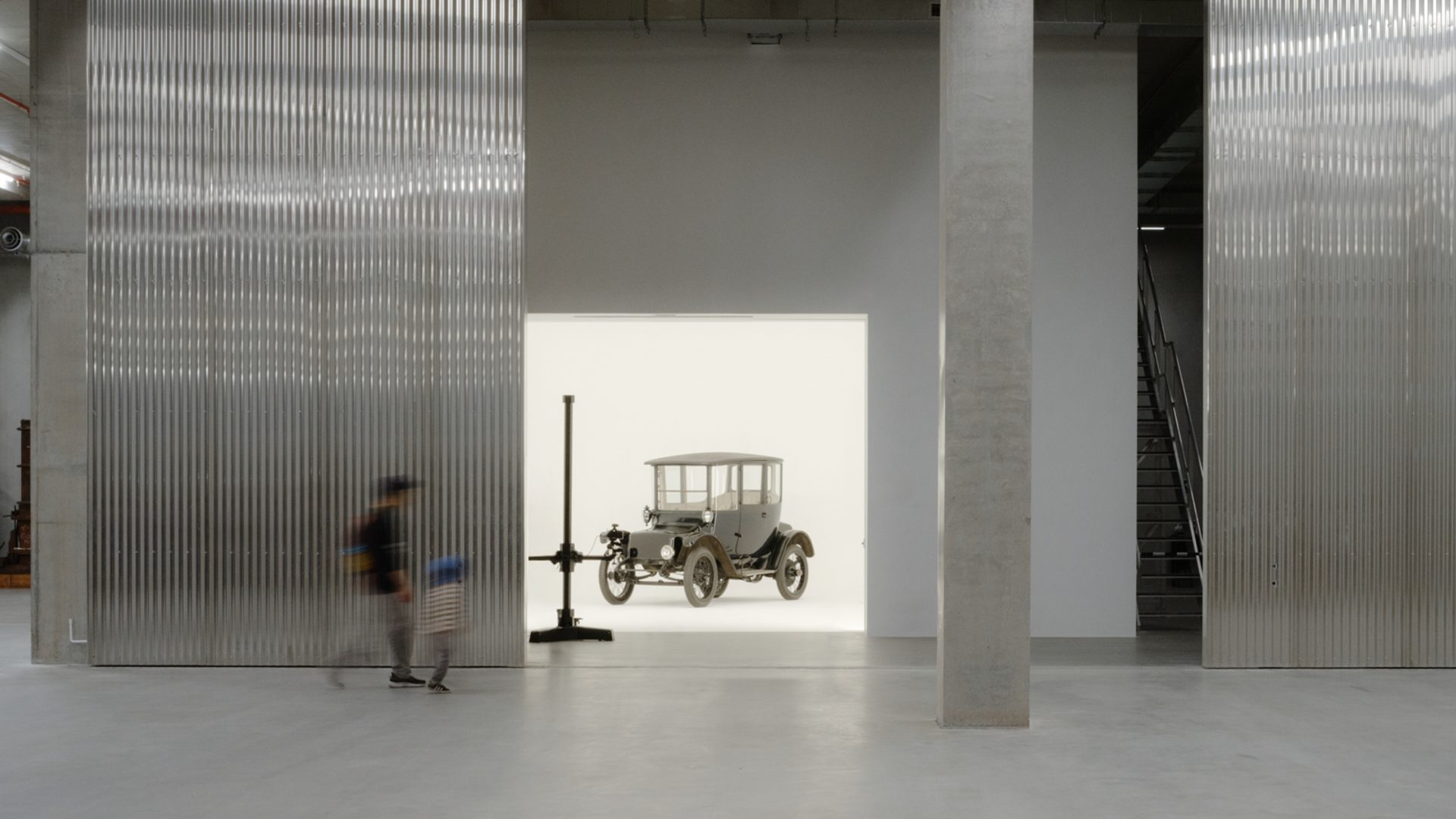A tall and shorter human figures walk through the gigantic storage house, to find a car behind the wall in a white room.