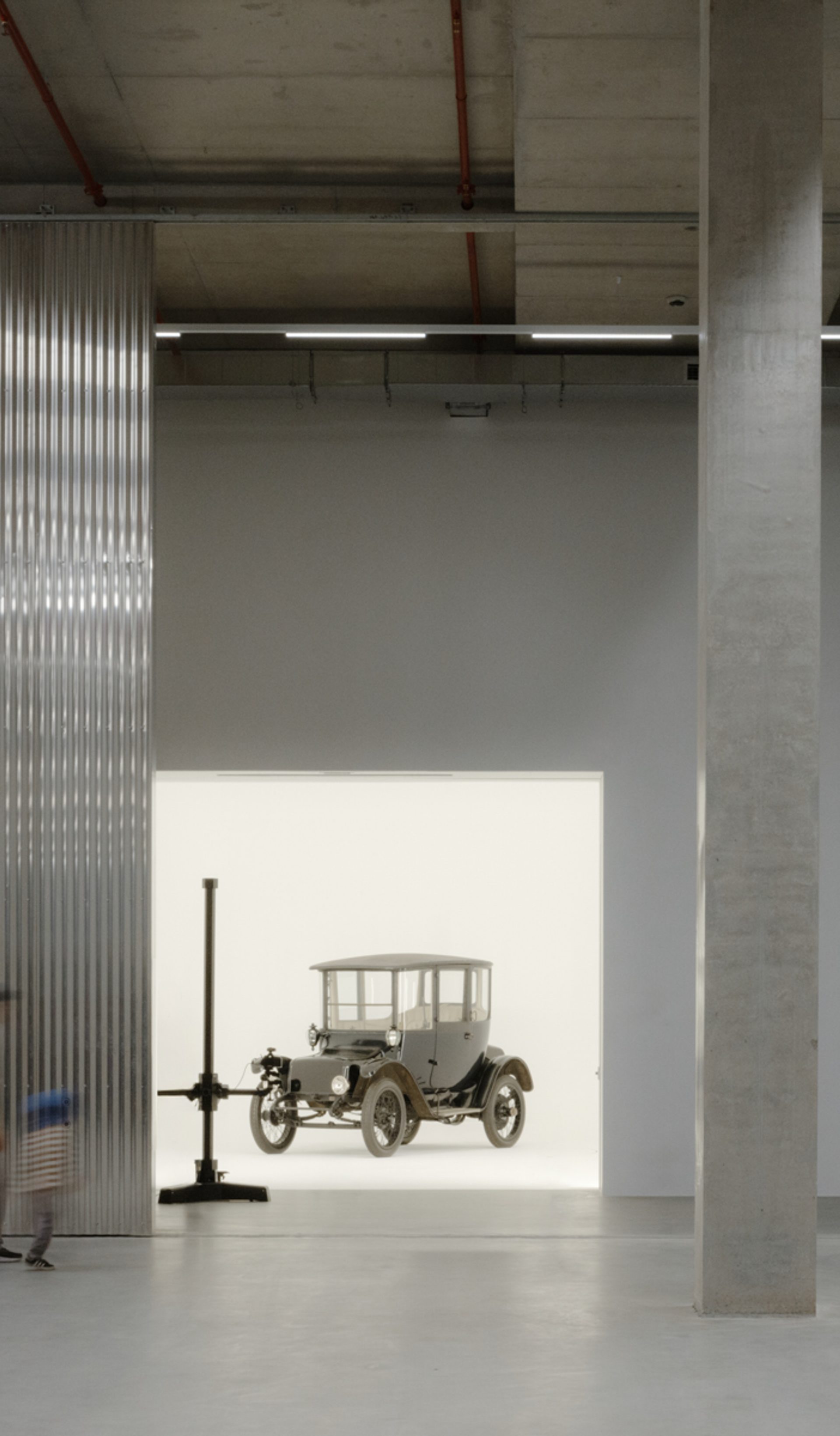 A tall and shorter human figures walk through the gigantic storage house, to find a car behind the wall in a white room.