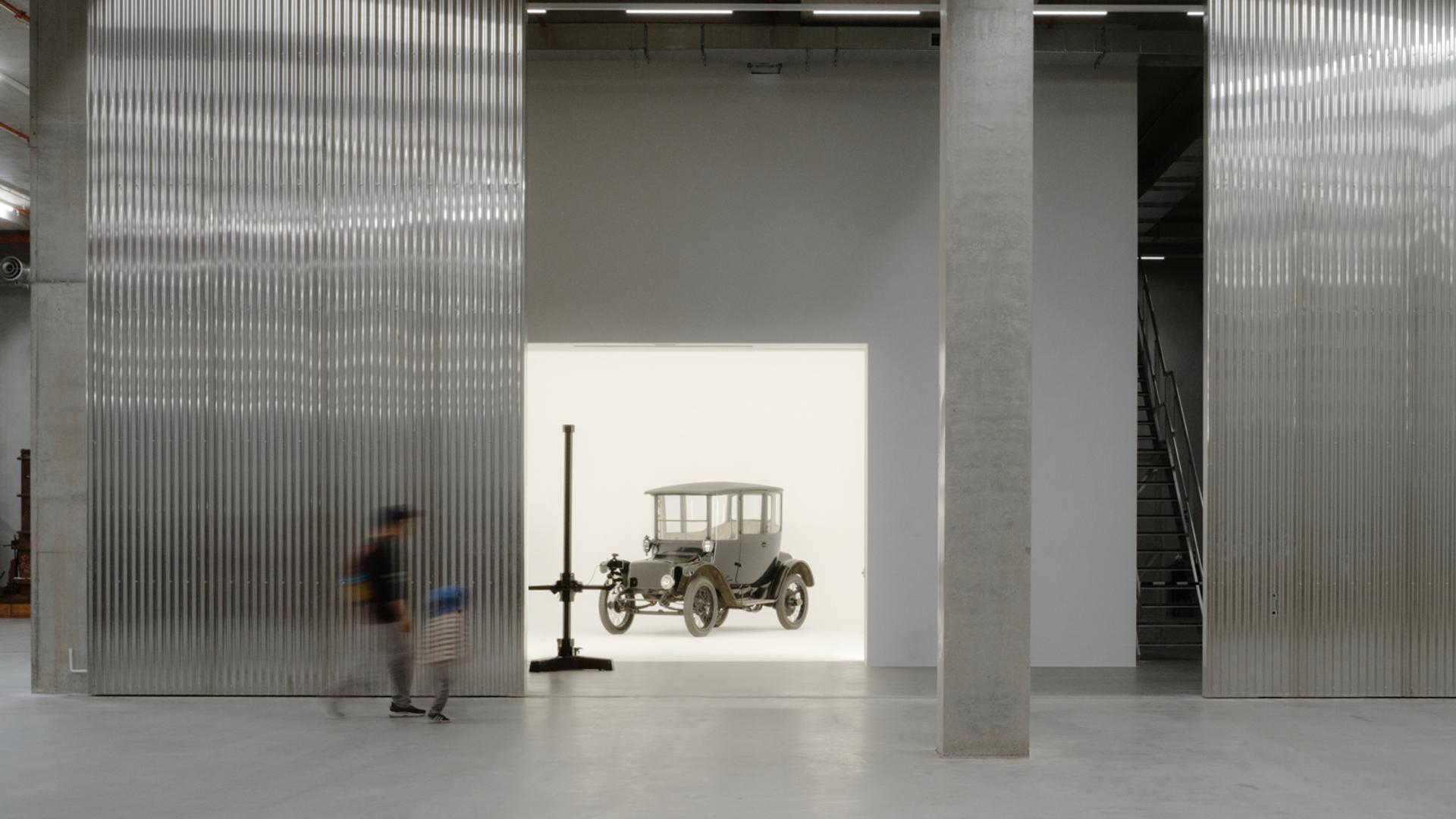 A tall and shorter human figures walk through the gigantic storage house, to find a car behind the wall in a white room.