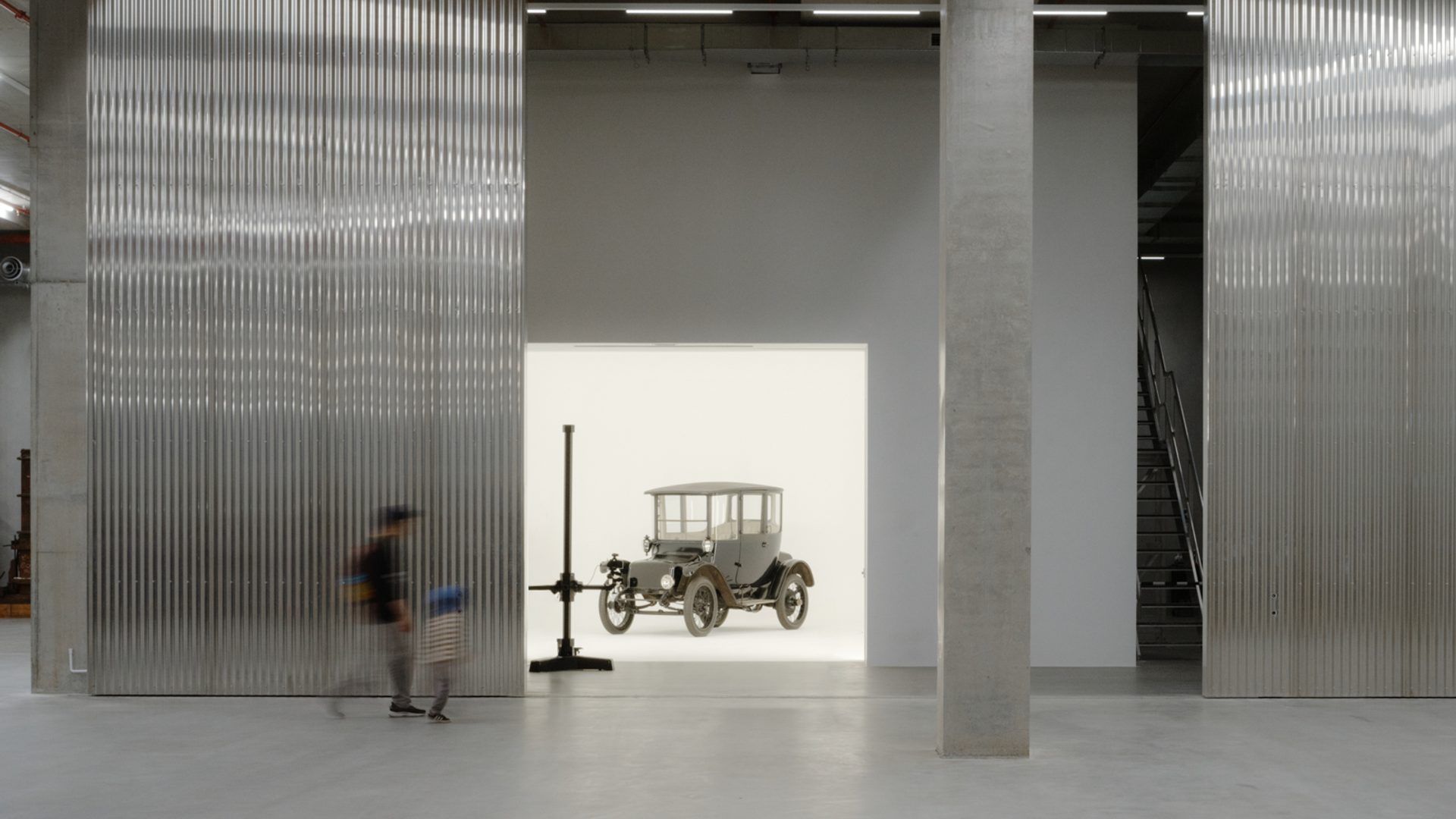 A tall and shorter human figures walk through the gigantic storage house, to find a car behind the wall in a white room.