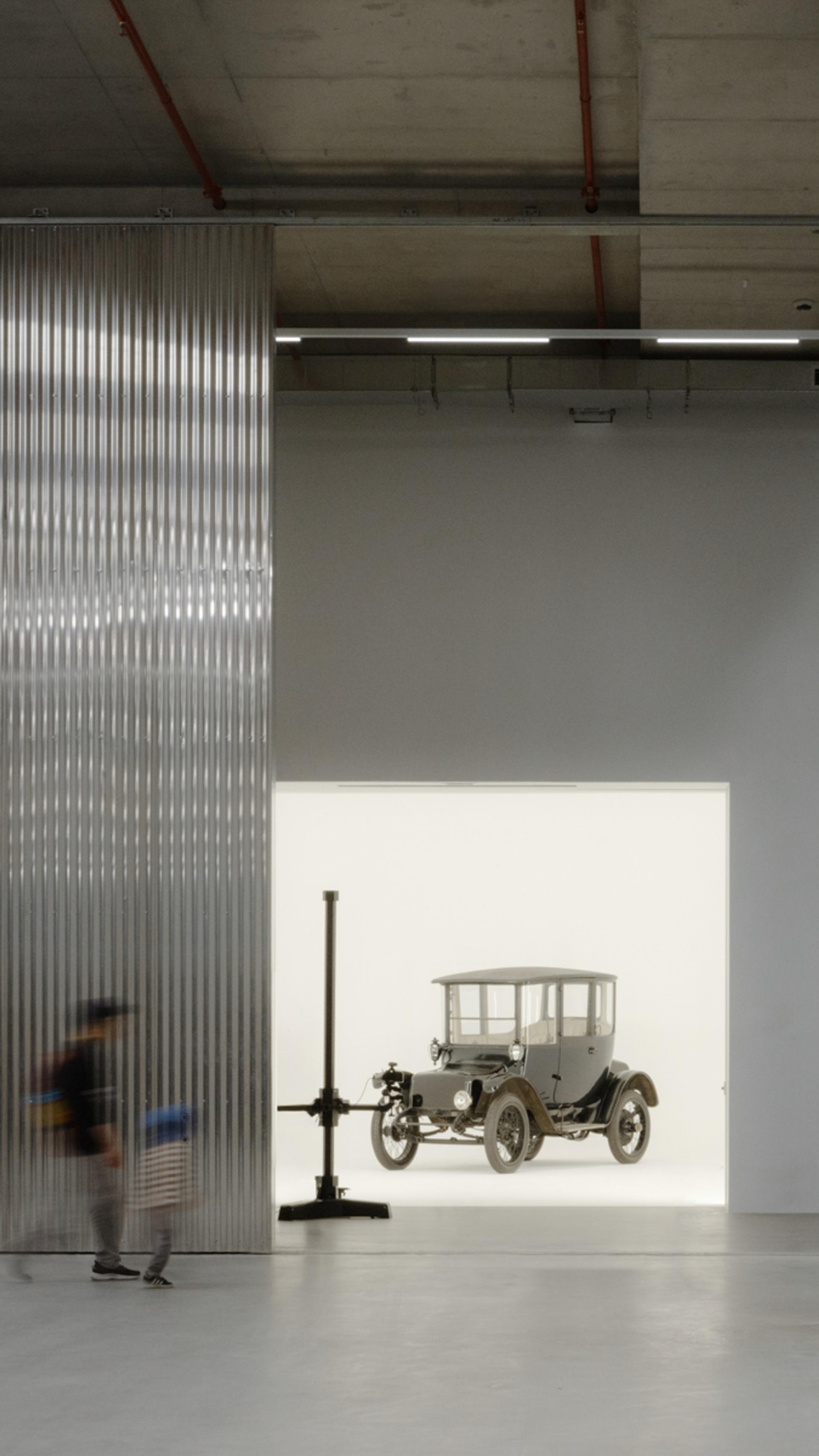 A tall and shorter human figures walk through the gigantic storage house, to find a car behind the wall in a white room.