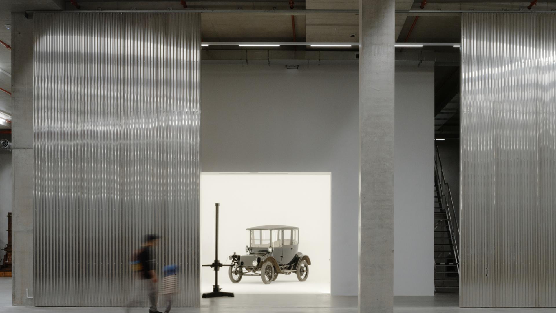 A tall and shorter human figures walk through the gigantic storage house, to find a car behind the wall in a white room.