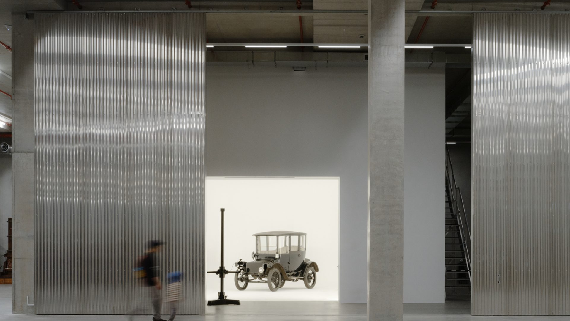 A tall and shorter human figures walk through the gigantic storage house, to find a car behind the wall in a white room.