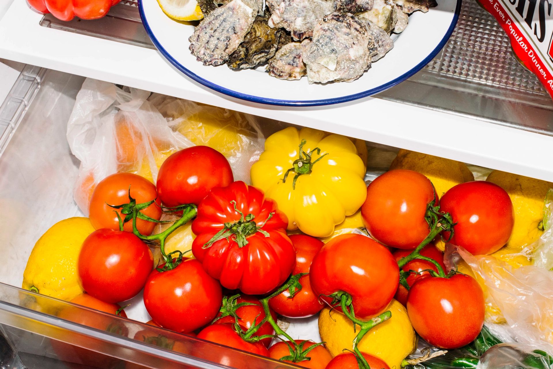 The vegetable drawer of a fridge is slightly open. The drawer is full of red and yellow tomatoes.