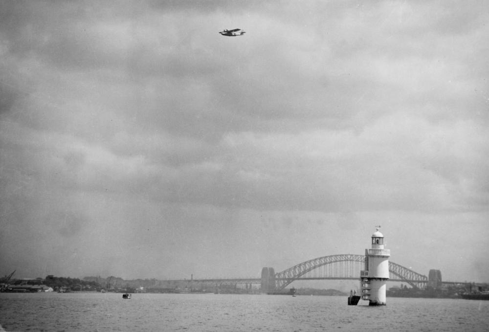 The Catalina flies above the Sydney Harbour Bridge. Black and white photograph.