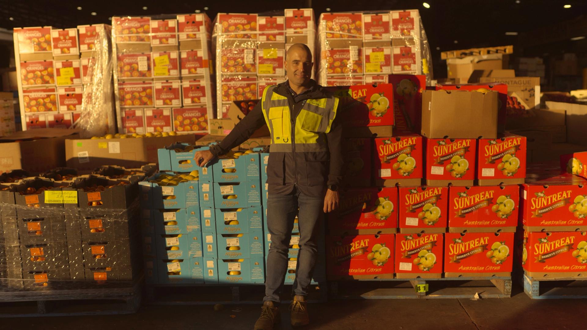 A man wearing a high-visibility vest stands in a large warehouse filled with stacks of brightly colored boxes labeled with various fruit brands, including 'Suntreat' and 'Nippy's.' He leans against a pallet of blue boxes, smiling slightly as he poses for the camera. The warehouse is dimly lit, with light shining on the boxes, creating a warm, industrial atmosphere.