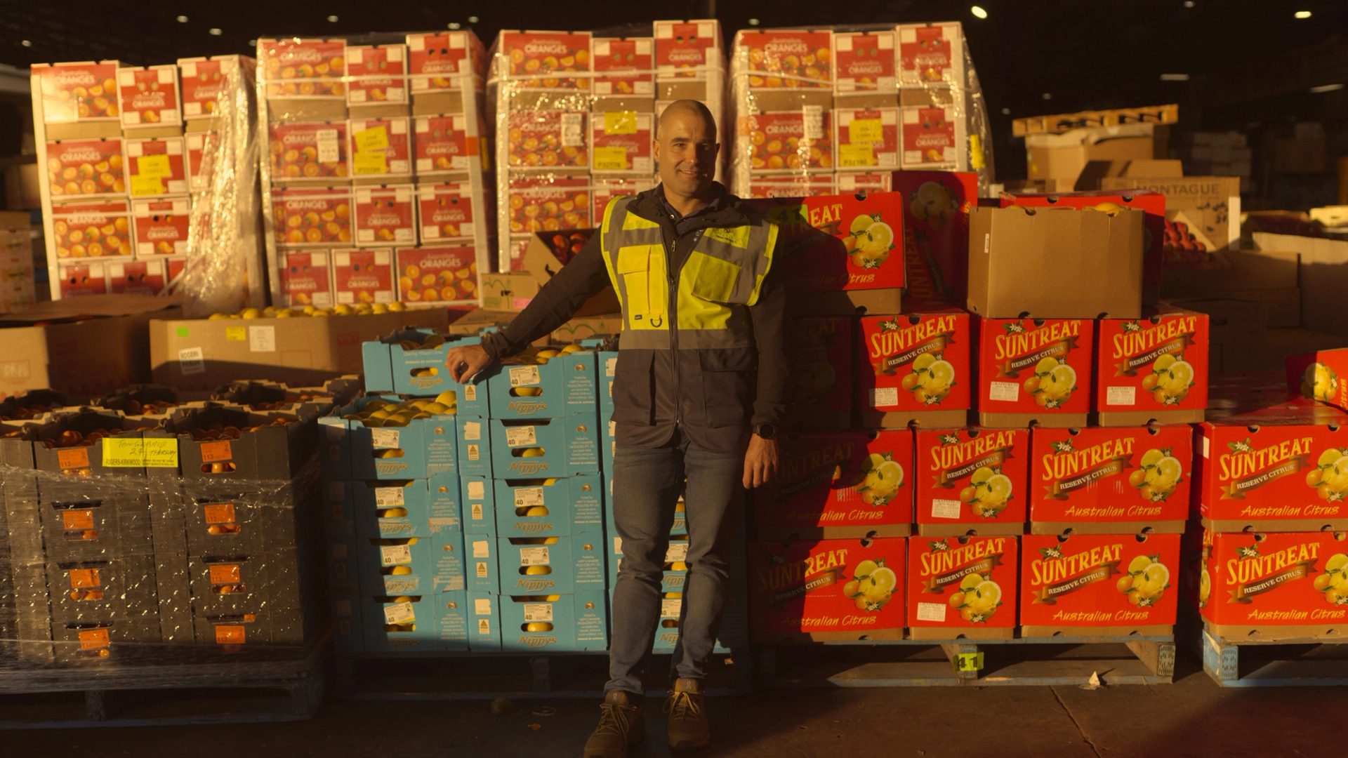 A man wearing a high-visibility vest stands in a large warehouse filled with stacks of brightly colored boxes labeled with various fruit brands, including 'Suntreat' and 'Nippy's.' He leans against a pallet of blue boxes, smiling slightly as he poses for the camera. The warehouse is dimly lit, with light shining on the boxes, creating a warm, industrial atmosphere.