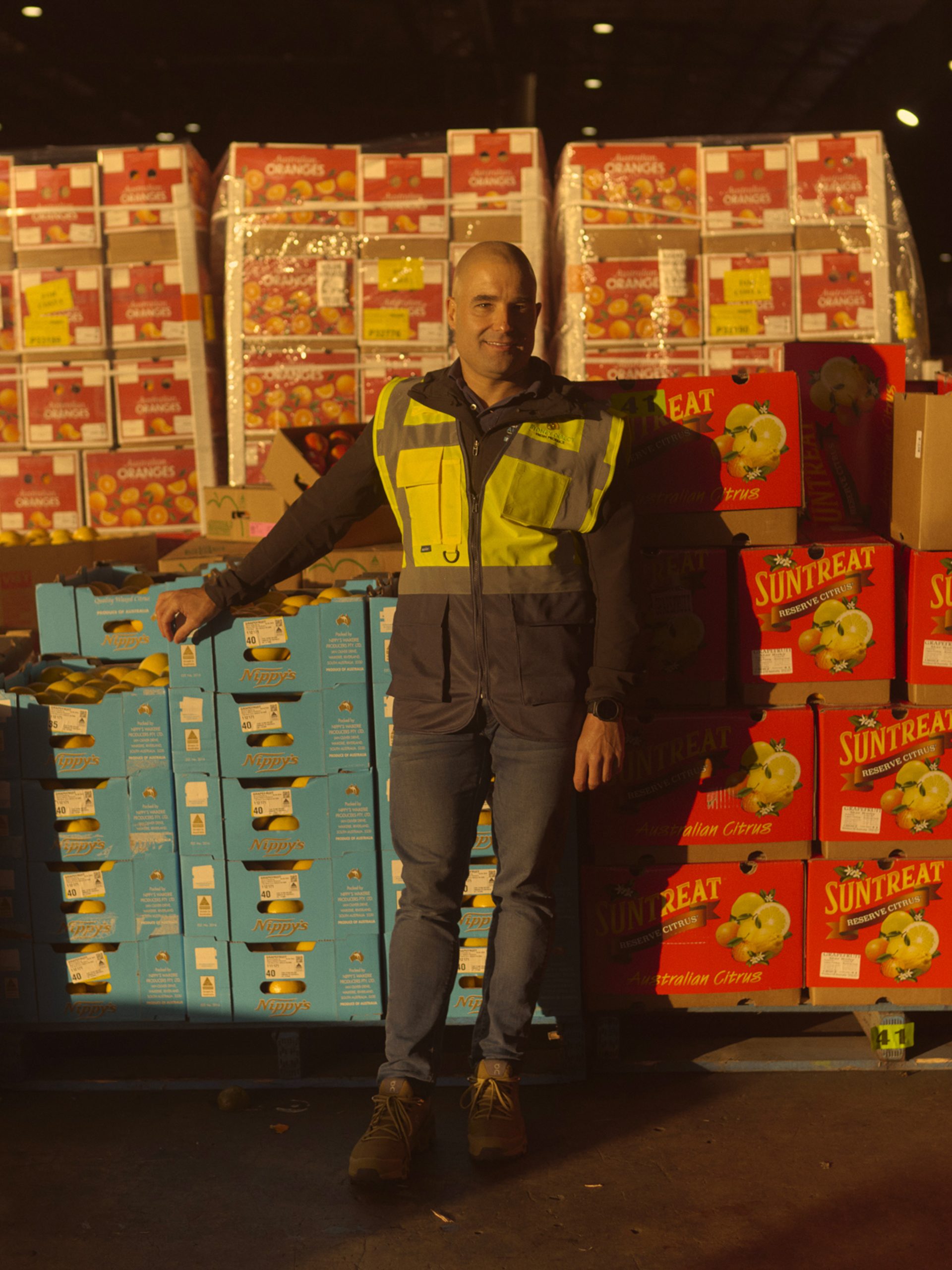 A man wearing a high-visibility vest stands in a large warehouse filled with stacks of brightly colored boxes labeled with various fruit brands, including 'Suntreat' and 'Nippy's.' He leans against a pallet of blue boxes, smiling slightly as he poses for the camera. The warehouse is dimly lit, with light shining on the boxes, creating a warm, industrial atmosphere.