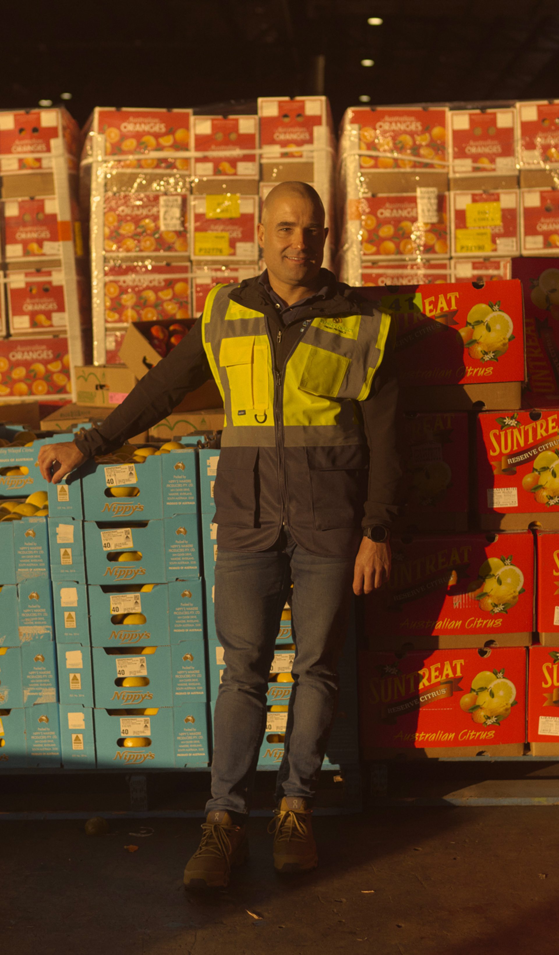 A man wearing a high-visibility vest stands in a large warehouse filled with stacks of brightly colored boxes labeled with various fruit brands, including 'Suntreat' and 'Nippy's.' He leans against a pallet of blue boxes, smiling slightly as he poses for the camera. The warehouse is dimly lit, with light shining on the boxes, creating a warm, industrial atmosphere.