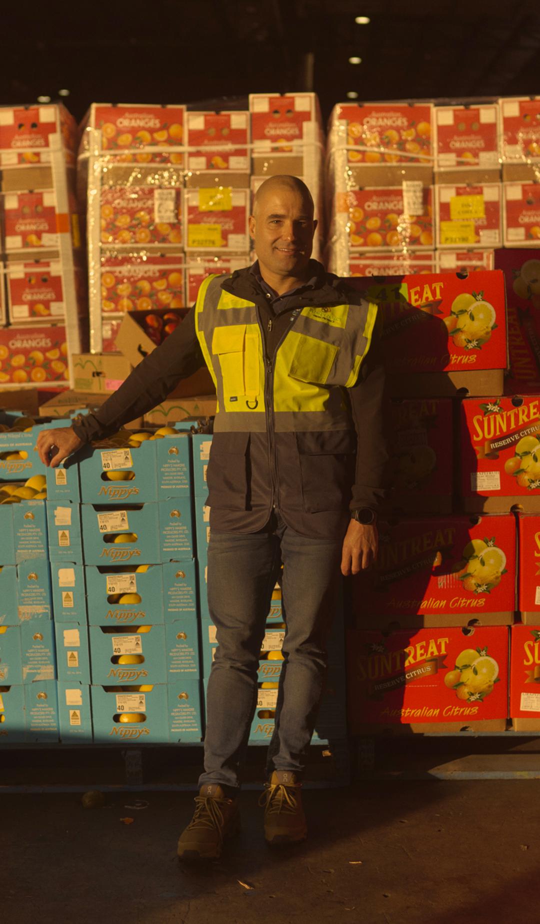 A man wearing a high-visibility vest stands in a large warehouse filled with stacks of brightly colored boxes labeled with various fruit brands, including 'Suntreat' and 'Nippy's.' He leans against a pallet of blue boxes, smiling slightly as he poses for the camera. The warehouse is dimly lit, with light shining on the boxes, creating a warm, industrial atmosphere.