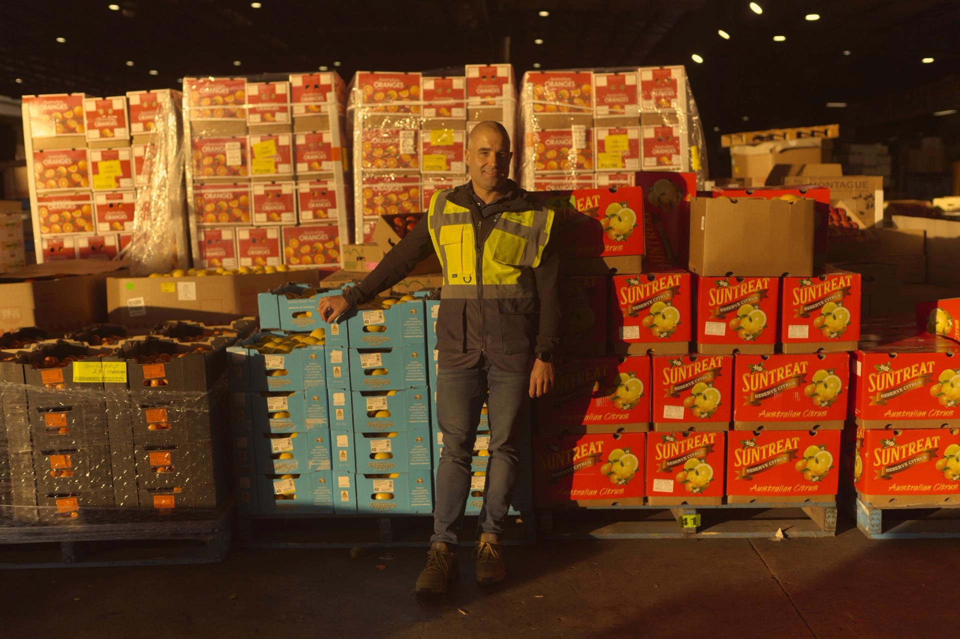 A man wearing a high-visibility vest stands in a large warehouse filled with stacks of brightly colored boxes labeled with various fruit brands, including 'Suntreat' and 'Nippy's.' He leans against a pallet of blue boxes, smiling slightly as he poses for the camera. The warehouse is dimly lit, with light shining on the boxes, creating a warm, industrial atmosphere.