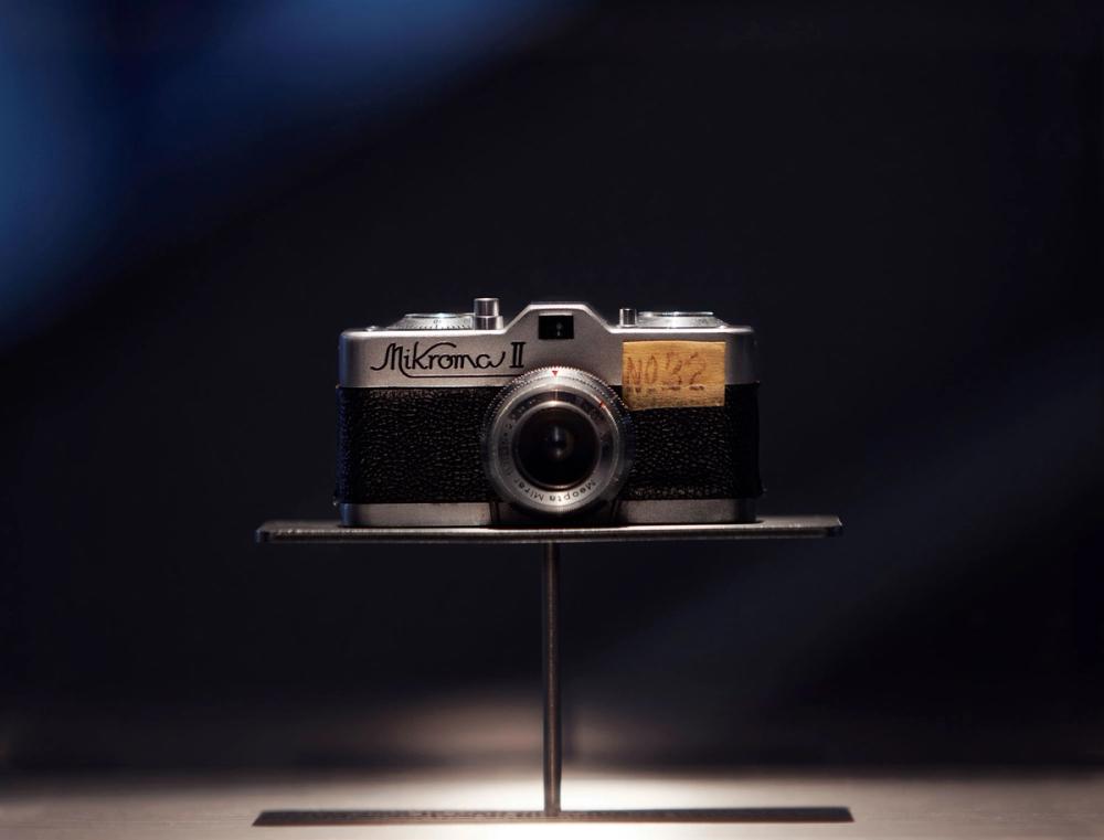 An exhibition view of a silver and black film camera on a metal platform.The shutter dials and counter are on the top. The glass lens have various dials around it and the view finder is on top.
