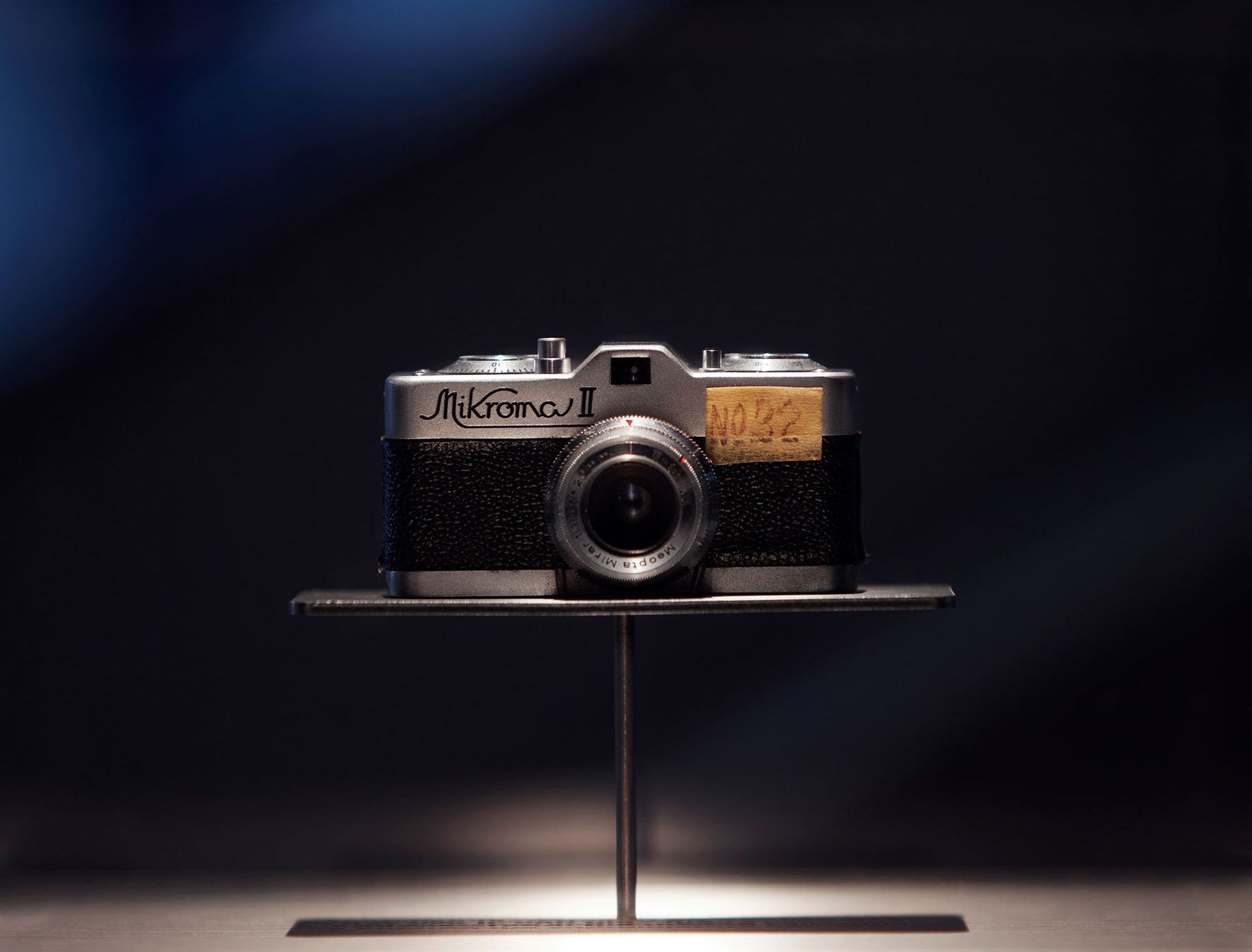 An exhibition view of a silver and black film camera on a metal platform.The shutter dials and counter are on the top. The glass lens have various dials around it and the view finder is on top.