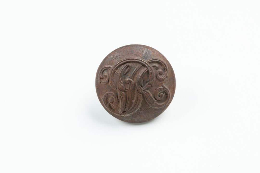 Circular metal button with embossed letters V R (Victoria Regina) intertwined in ornamental script on front surface. Button is concave, with inserted disc with loop on reverse for attaching to garment.