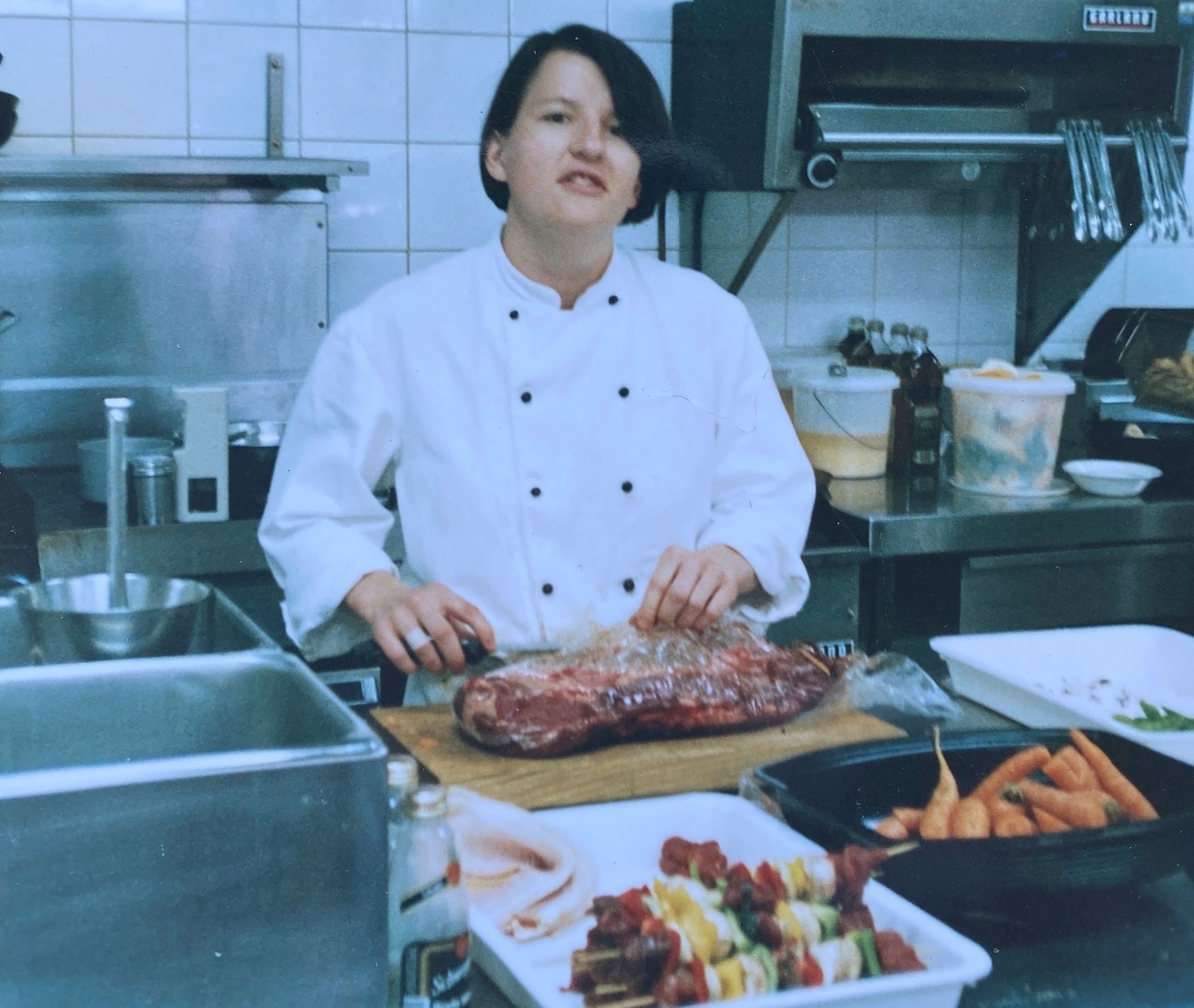 Chef in industrial kitchen, with a slab of meat on the chopping board, kebabs and carrots in front of her.