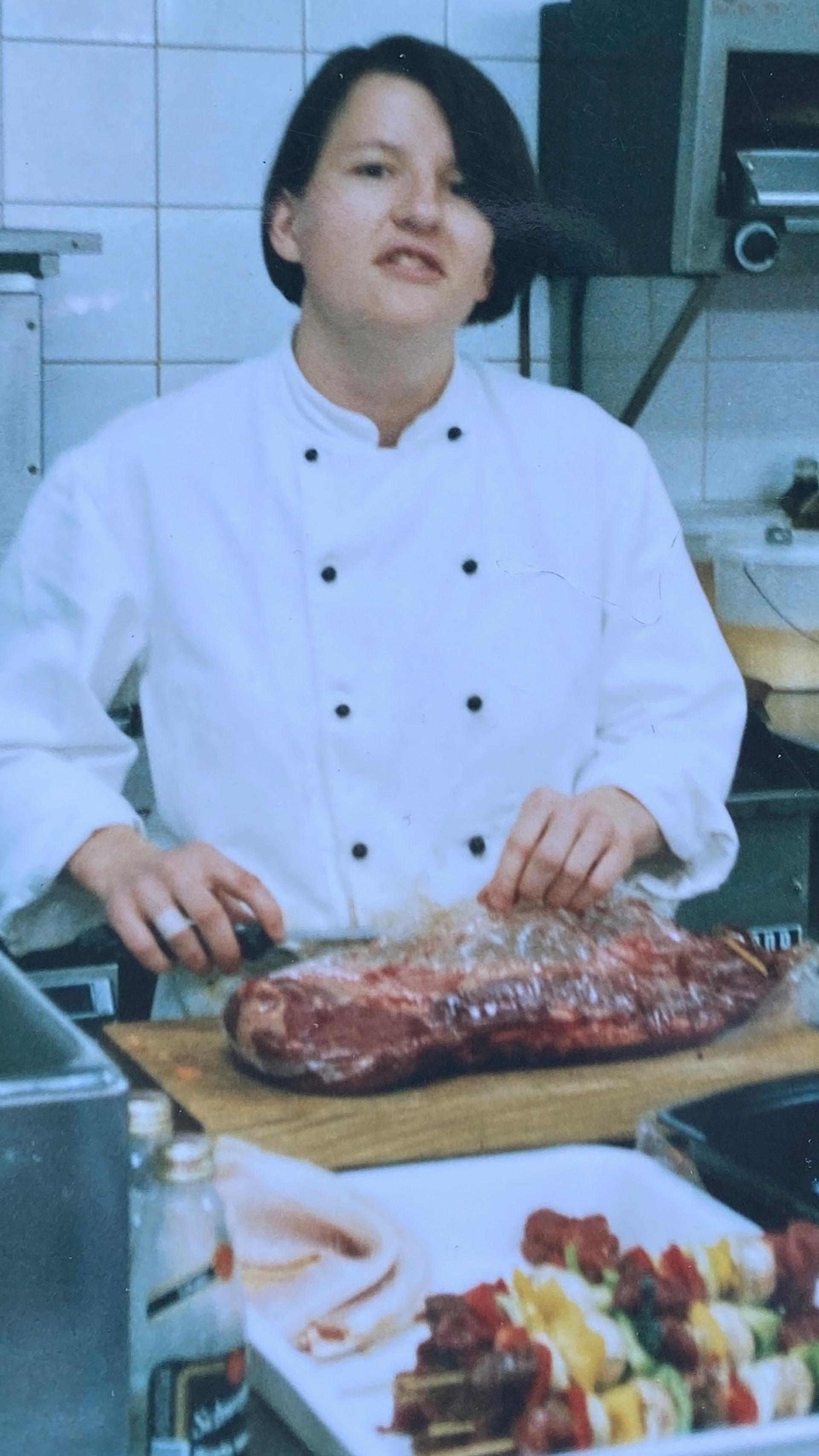 Chef in industrial kitchen, with a slab of meat on the chopping board, kebabs and carrots in front of her.