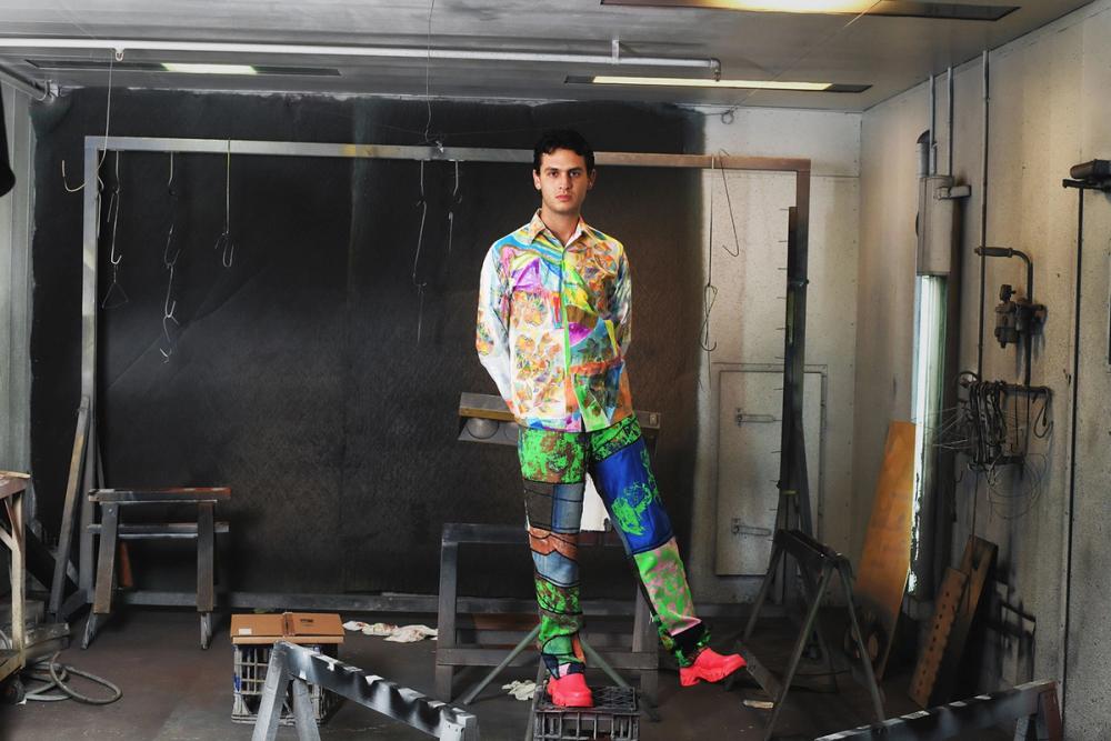 Jordan Gogos stand on a milk crate in the Powerhouse workshop. He wears a multicoloured shirt and pants with bright red shoes.