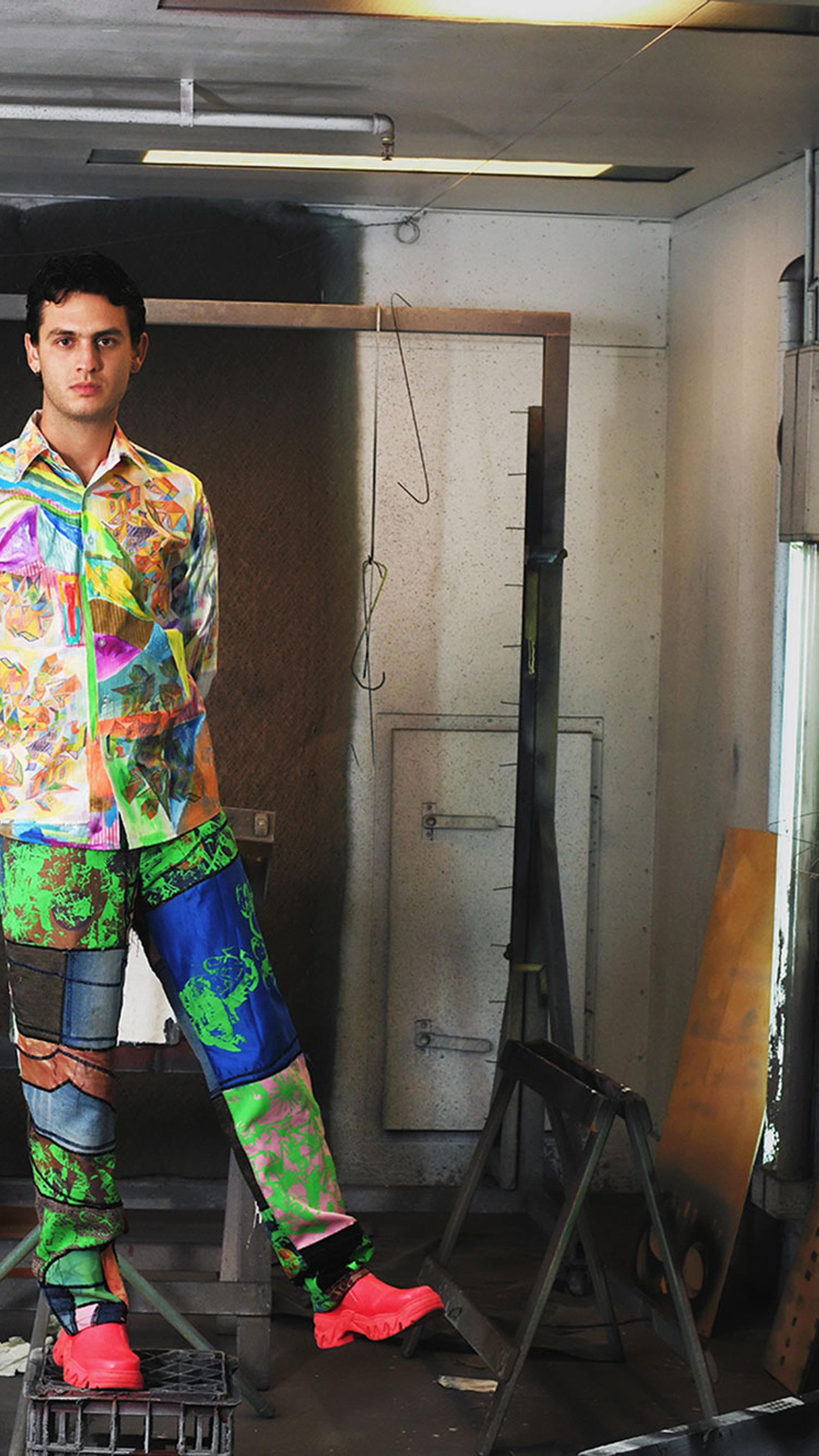 Jordan Gogos stand on a milk crate in the Powerhouse workshop. He wears a multicoloured shirt and pants with bright red shoes.