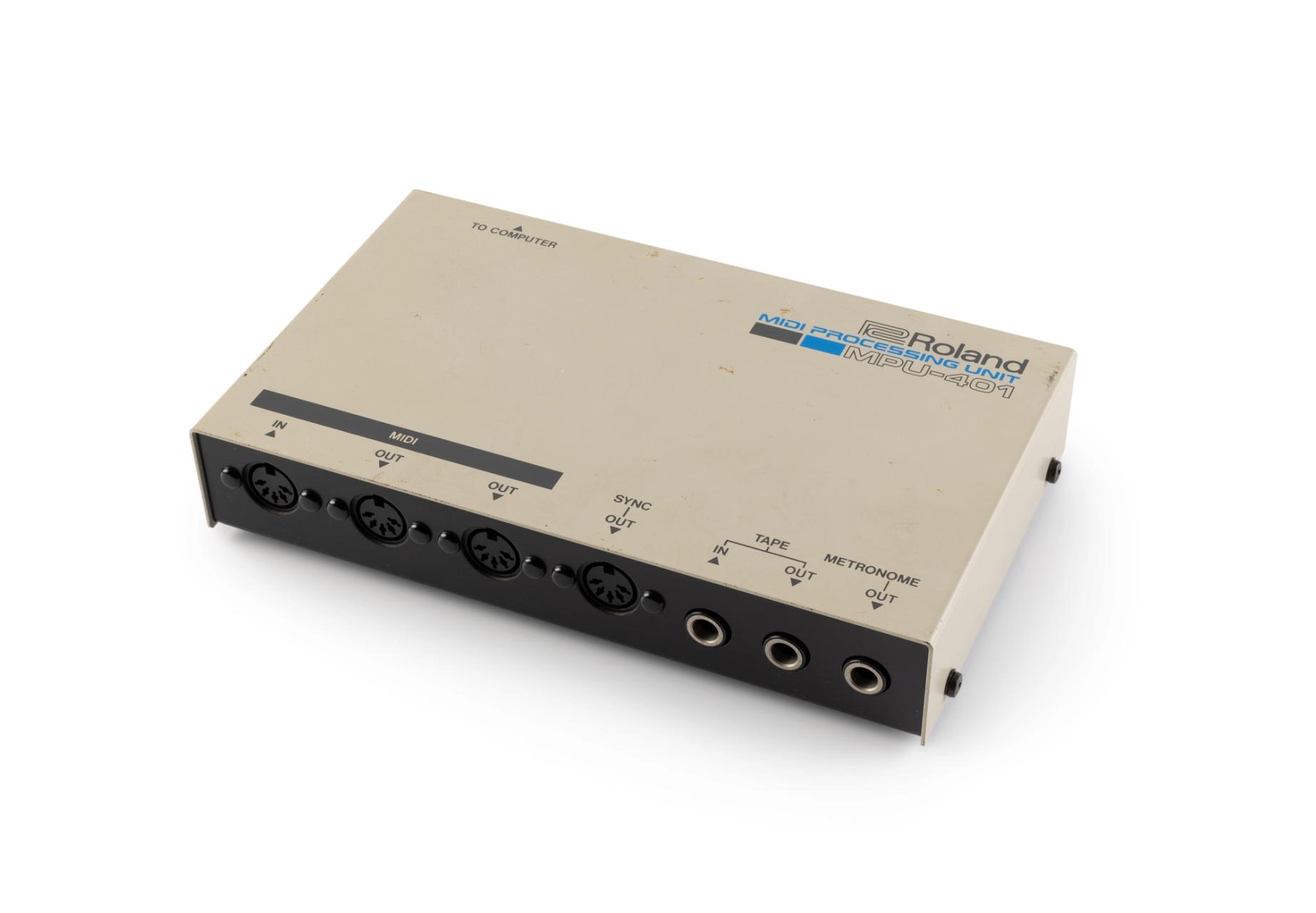 A Roland MIDI Processing Unit MPU-401 consisting of black coloured rectangular metal box covered in a casing finished in pale grey. One side of the unit has port for attachment to computer, the other side has ports for MIDI, Sync, Tape and Metronome. The manufacturer details are printed on the top right hand corner and there are details printed on labels attached to the base of the unit.
