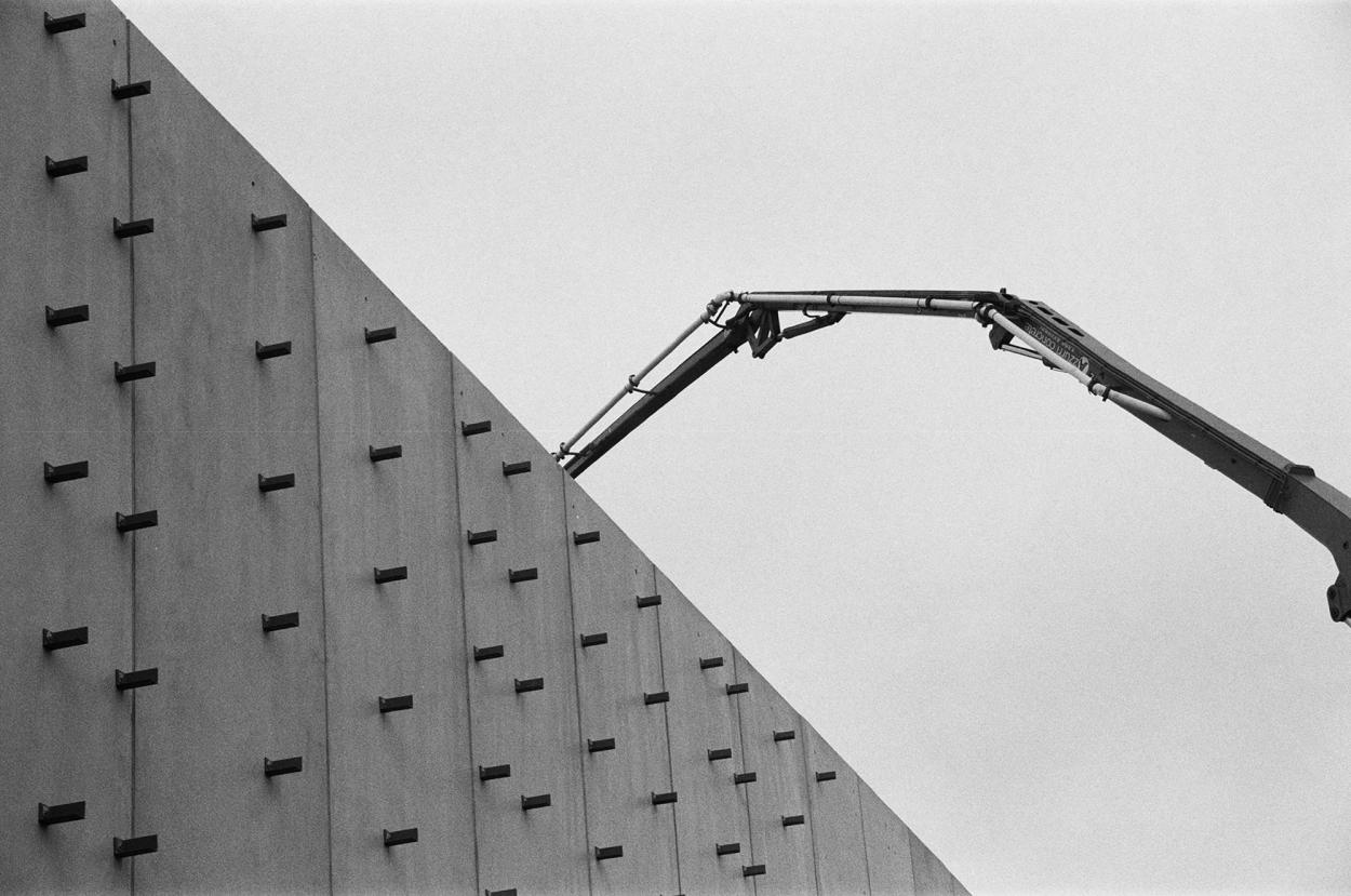 A black and white photograph of a crane’s arm reaching over a concrete wall.