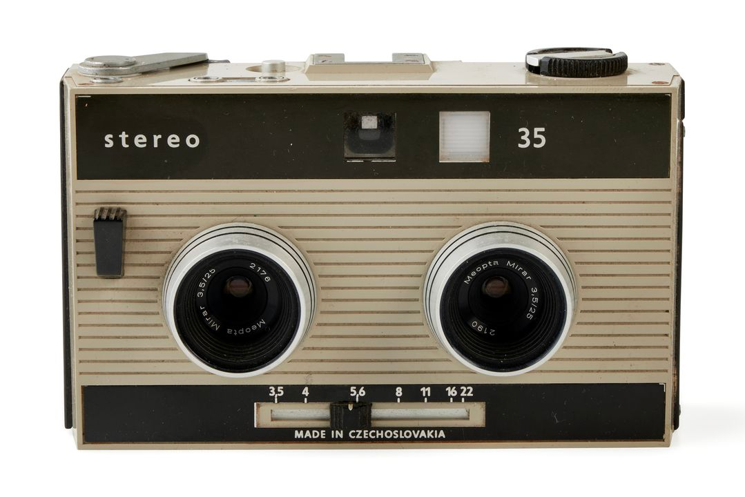 The Meopta 35 stereo camera consists of a grey plastic casing with black trim and 'stereo / 35' on the front. It features two lenses at the front, with an adjustment bar below for altering the aperture or exposure, and a square view finder above. The shutter button is positioned at the front, to one side of the lenses. The top of the device has a film winder and rewinder, one on each side and a metal clip in the centre for attachments.