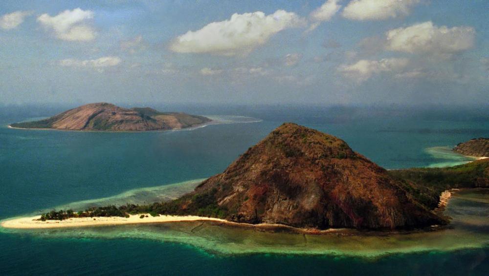 Colour photograph of two small hilly islands, the nearest one has a sand spit on the left side and is surrounded by light turquoise water.