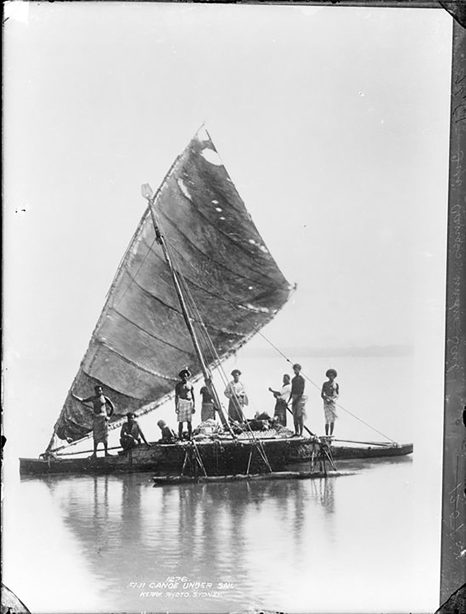An outrigger canoe with a large sail on the water. Seven men can be seen standing and two sitting on the canoe.