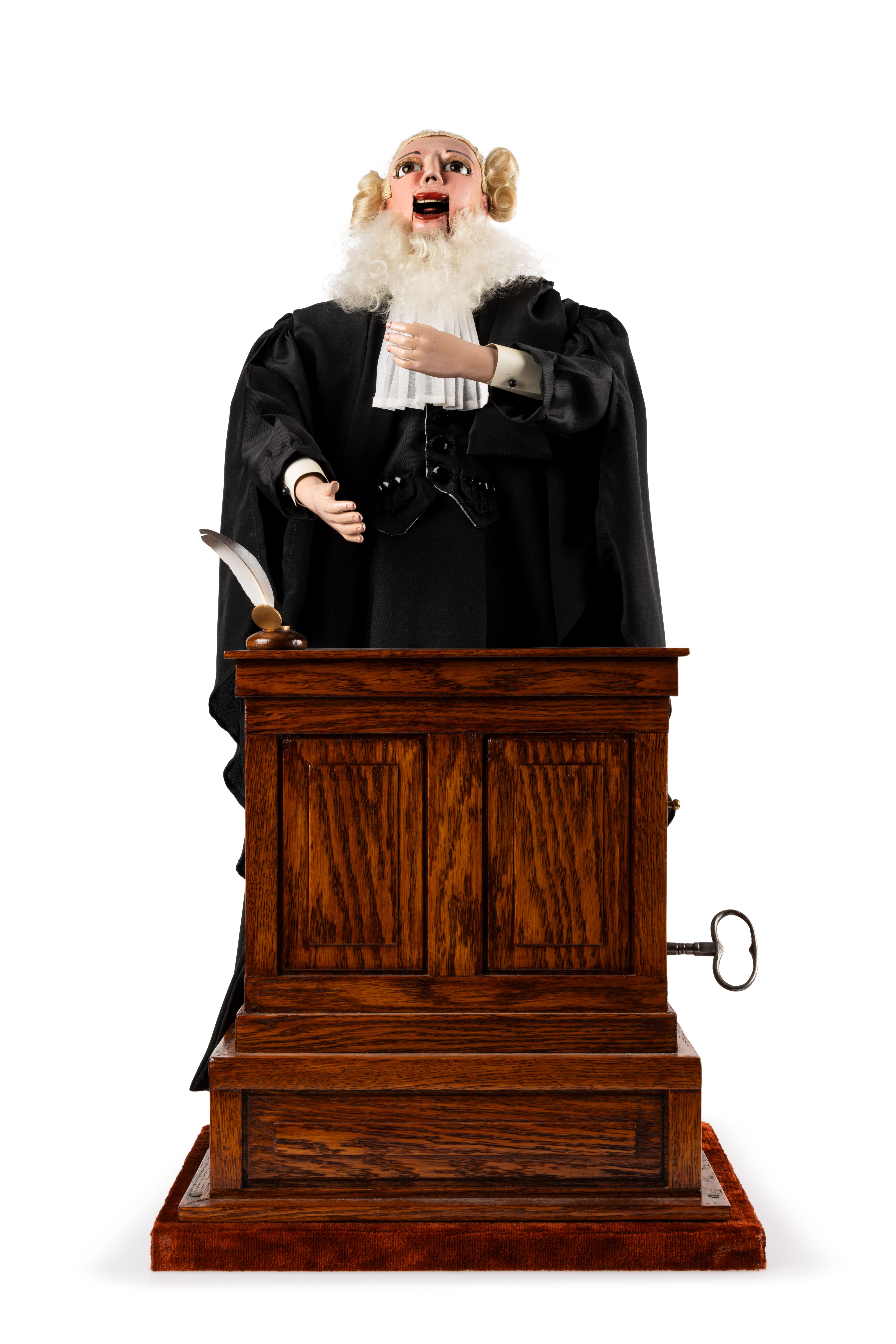 Musical automaton featuring a male lawyer in black robe and wig standing behind a podium. The right hand is stretched out, while the left hand holds a rolled document. The lawyer is automated, and the front part of his jaw is jointed so he could appear to speak. The mechanism, made up of wooden cogs and wires, is underneath the podium. A metal key for winding the mechanism protrudes from the side of the podium to the lawyer's left. The quill pen has broken off the inkwell on top of the podium and is tucked into the pocket of the lawyer's robe. The automaton plays a tune while the arms move up and down, the eyes blink and the mouth opens and closes.