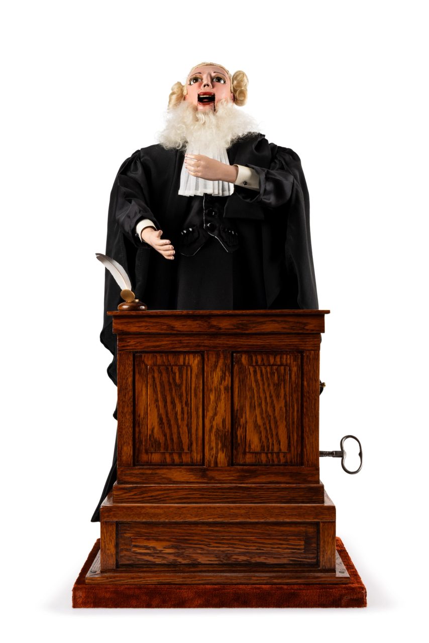 Musical automaton featuring a male lawyer in black robe and wig standing behind a podium. The right hand is stretched out, while the left hand holds a rolled document. The lawyer is automated, and the front part of his jaw is jointed so he could appear to speak. The mechanism, made up of wooden cogs and wires, is underneath the podium. A metal key for winding the mechanism protrudes from the side of the podium to the lawyer's left. The quill pen has broken off the inkwell on top of the podium and is tucked into the pocket of the lawyer's robe. The automaton plays a tune while the arms move up and down, the eyes blink and the mouth opens and closes.