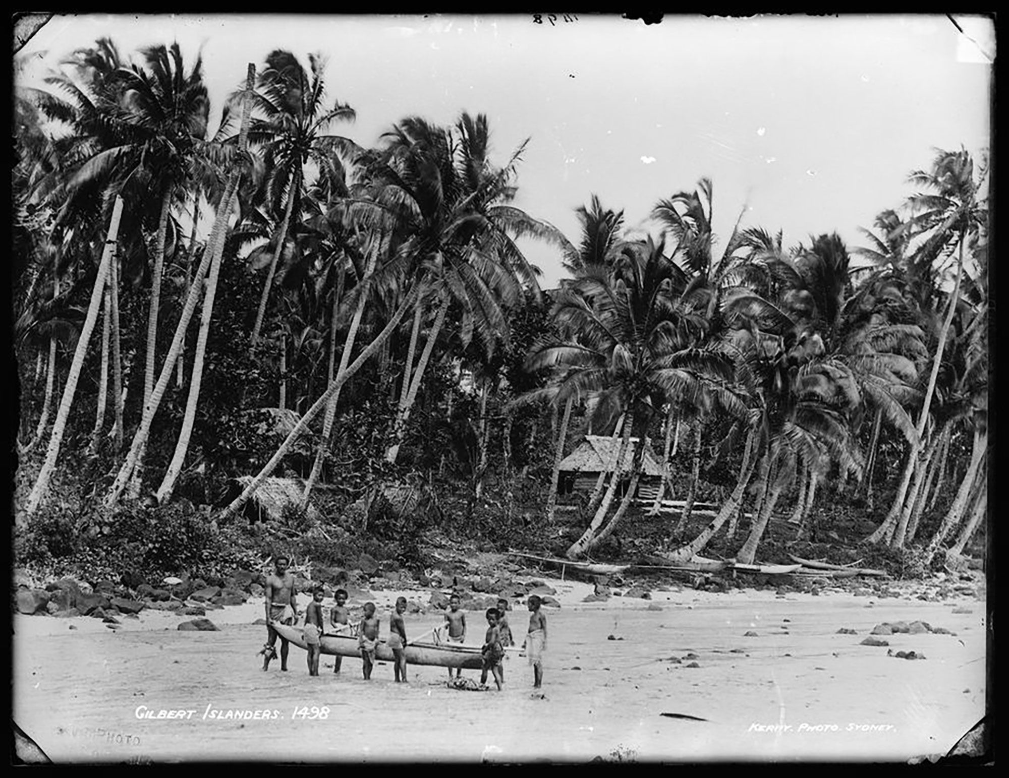 A man and a group of 8 boys carrying an outrigger canoe on a beach. Black and white photograph.