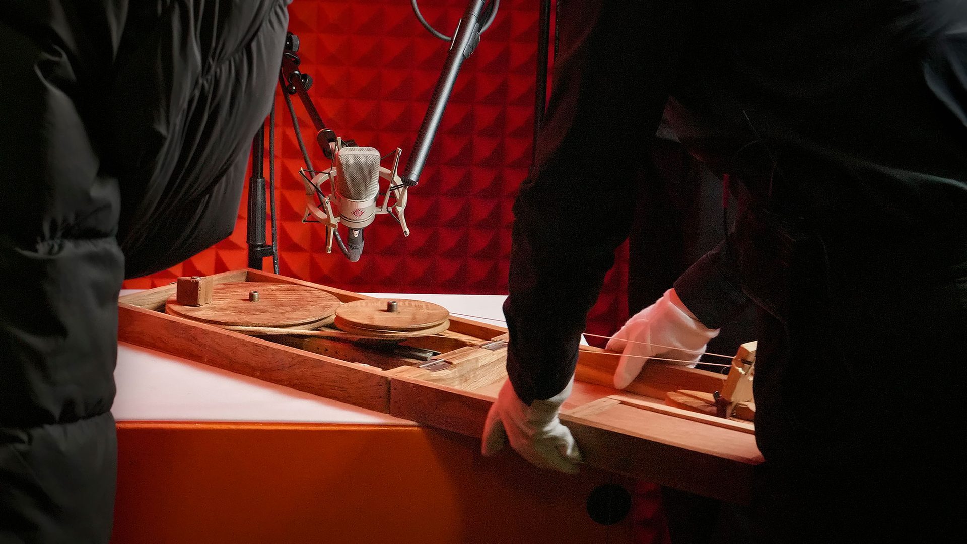 A wooden chakra (spinning wheel) sits on a box inside an orange sound booth. A silver microphone is placed above it and two figures stand either side with the figure on the right holding the chakra.