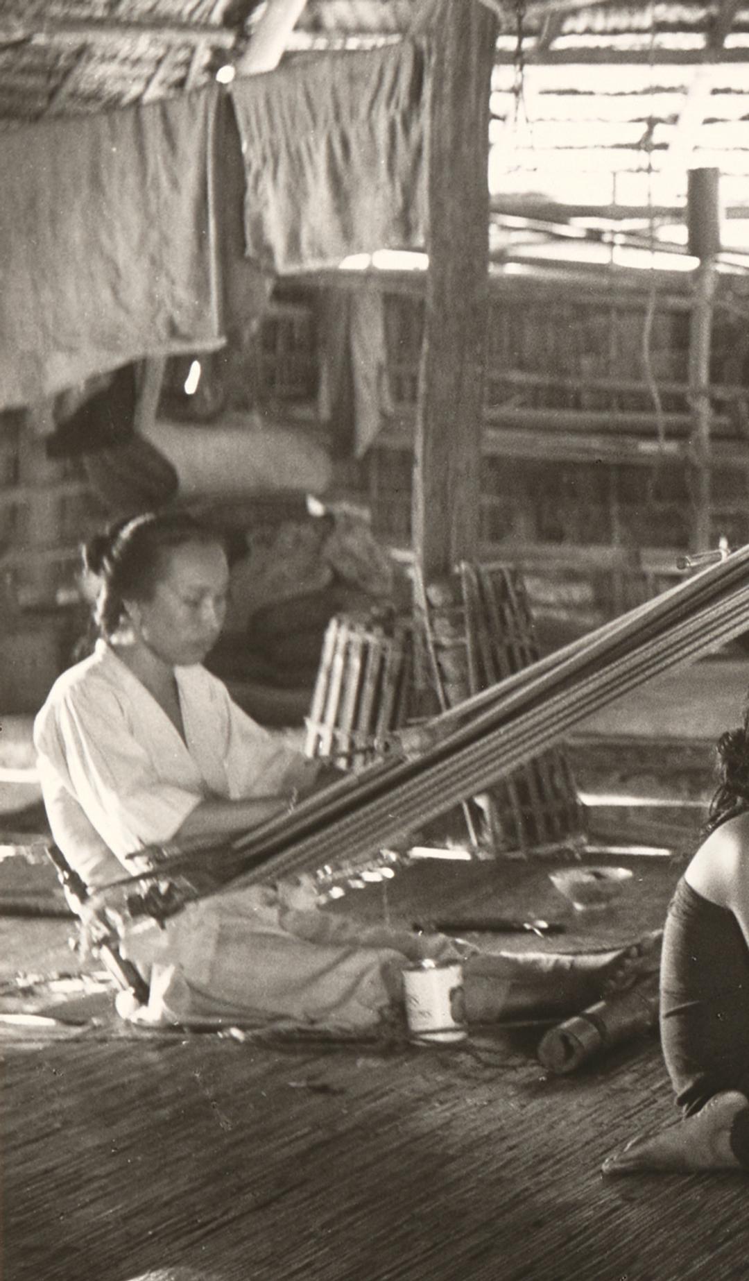 Black and white photograph showing two women, using back strap looms in a hut while two girls sit between them, in the centre of the photograph. The back strap loom used the weaver's body to provide the correct tension to the warps, the other end of which are tied to a rod, which is suspended from the roof timbers.