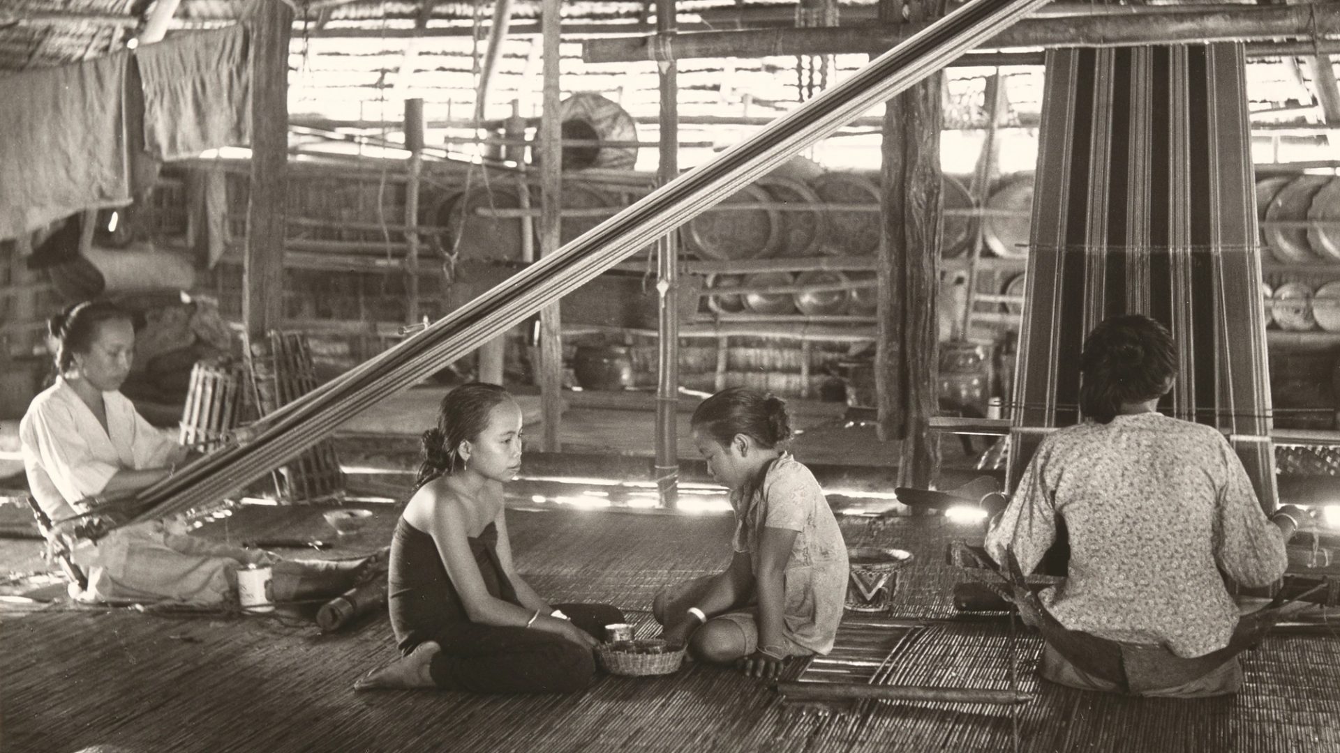 Black and white photograph showing two women, using back strap looms in a hut while two girls sit between them, in the centre of the photograph. The back strap loom used the weaver's body to provide the correct tension to the warps, the other end of which are tied to a rod, which is suspended from the roof timbers.