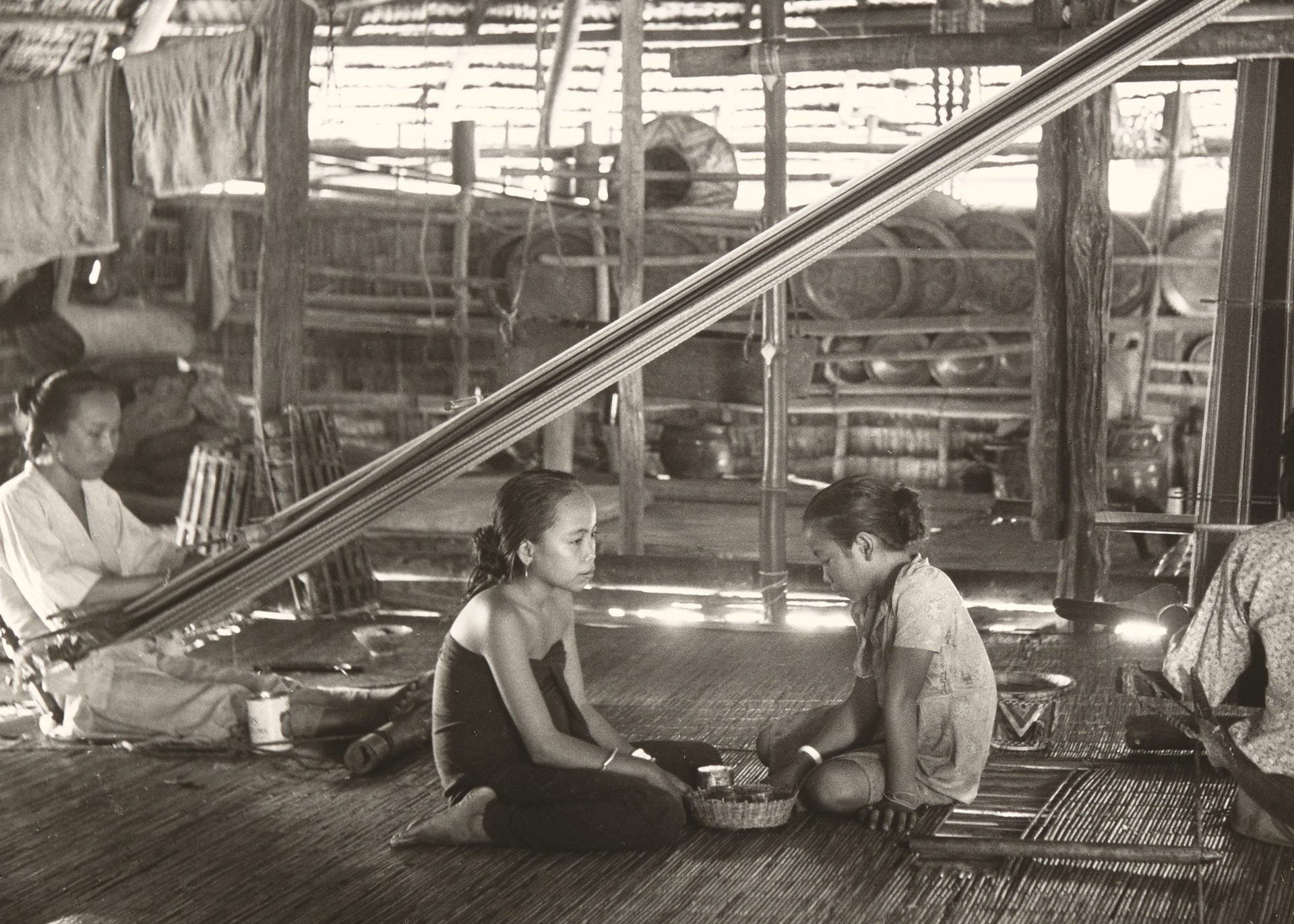 Black and white photograph showing two women, using back strap looms in a hut while two girls sit between them, in the centre of the photograph. The back strap loom used the weaver's body to provide the correct tension to the warps, the other end of which are tied to a rod, which is suspended from the roof timbers.