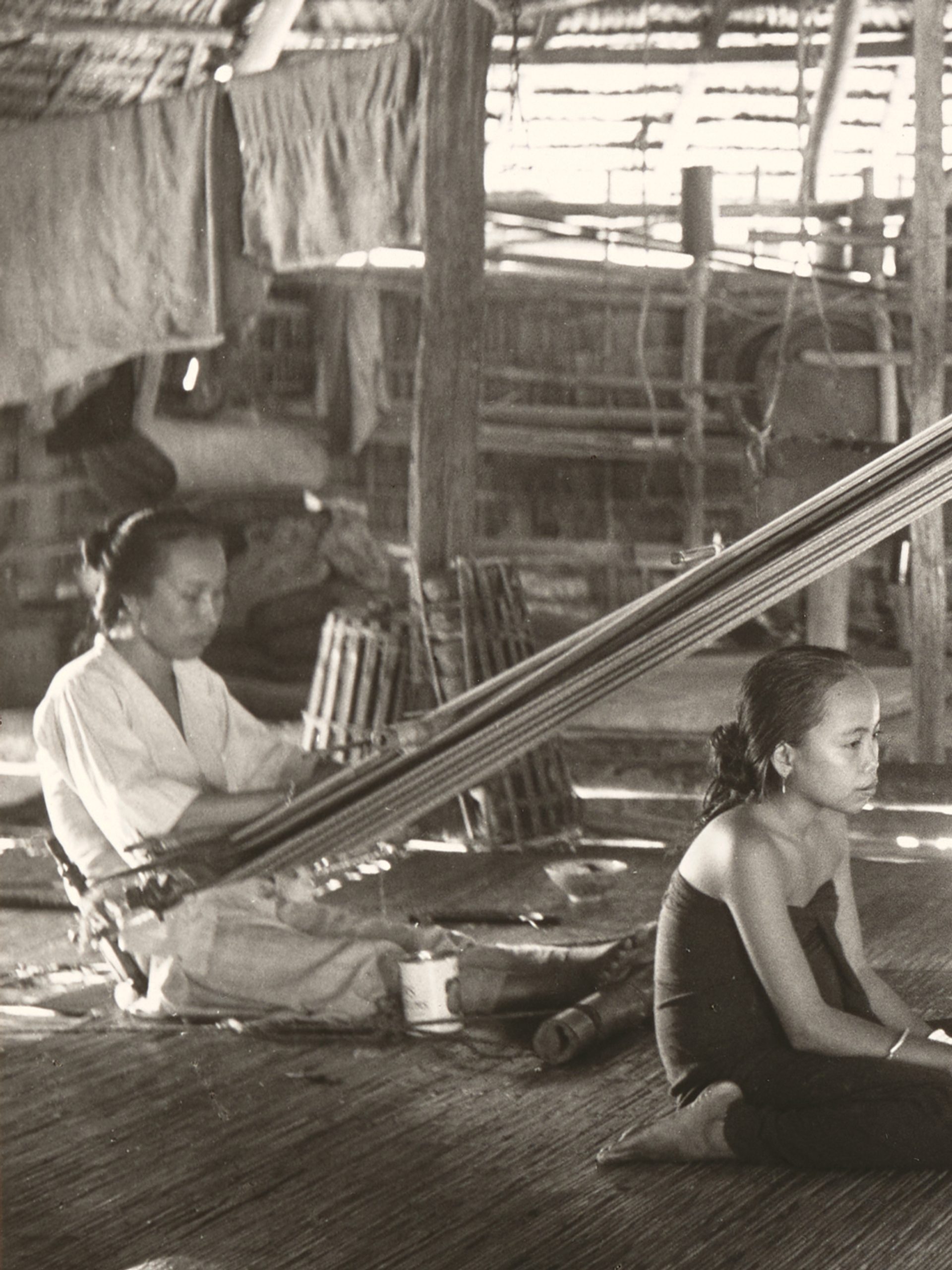 Black and white photograph showing two women, using back strap looms in a hut while two girls sit between them, in the centre of the photograph. The back strap loom used the weaver's body to provide the correct tension to the warps, the other end of which are tied to a rod, which is suspended from the roof timbers.