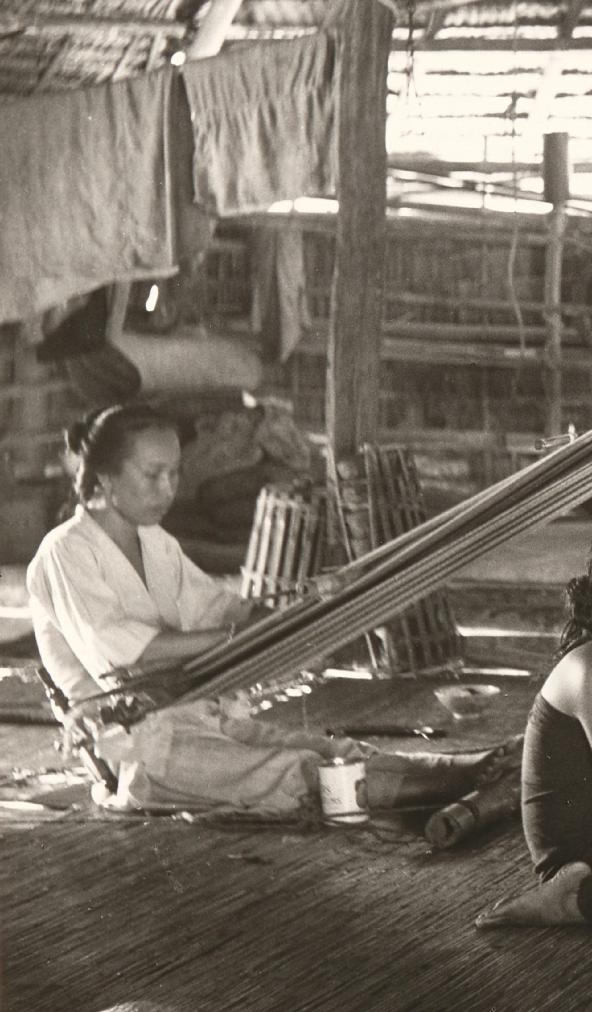 Black and white photograph showing two women, using back strap looms in a hut while two girls sit between them, in the centre of the photograph. The back strap loom used the weaver's body to provide the correct tension to the warps, the other end of which are tied to a rod, which is suspended from the roof timbers.