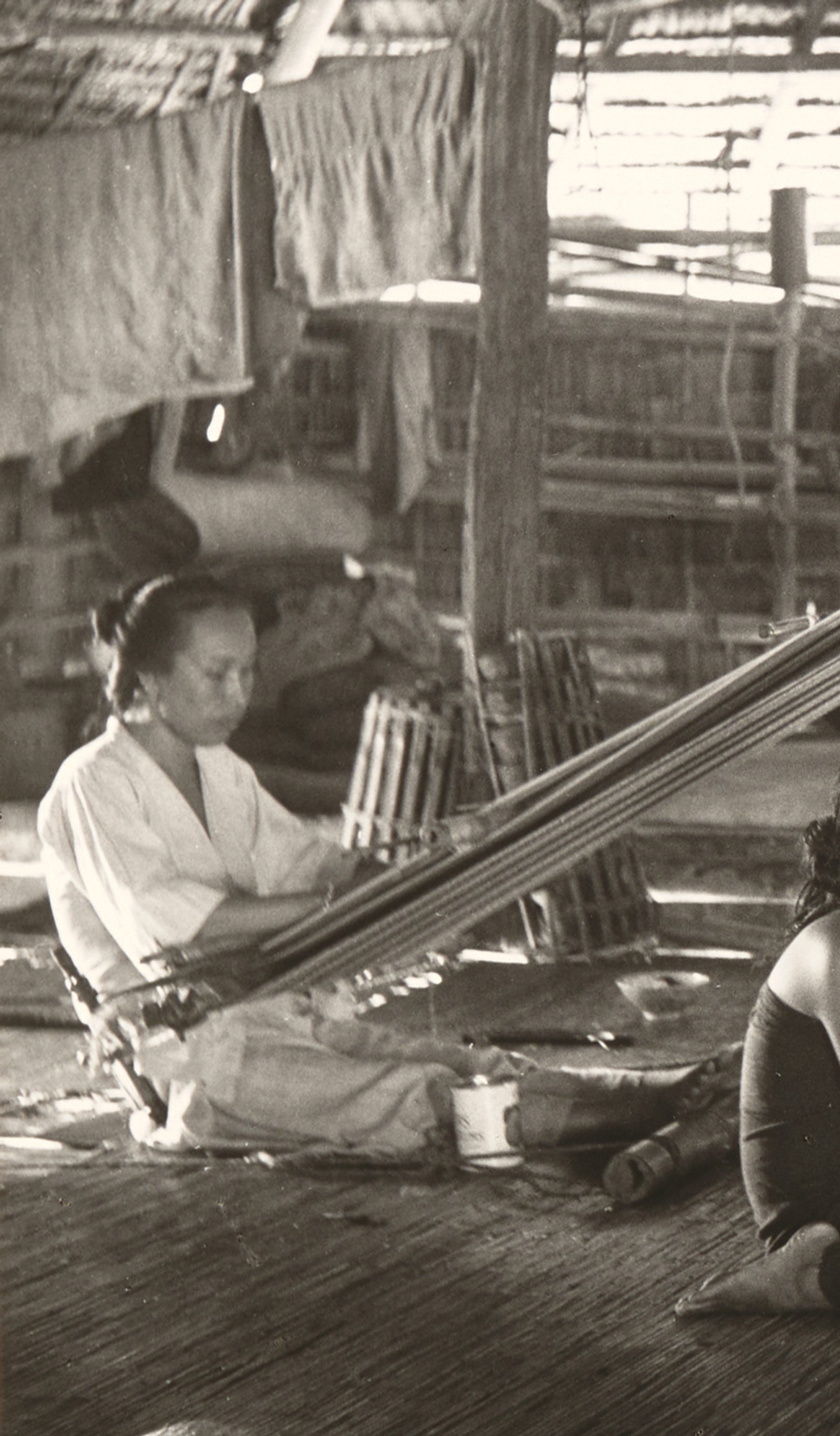 Black and white photograph showing two women, using back strap looms in a hut while two girls sit between them, in the centre of the photograph. The back strap loom used the weaver's body to provide the correct tension to the warps, the other end of which are tied to a rod, which is suspended from the roof timbers.